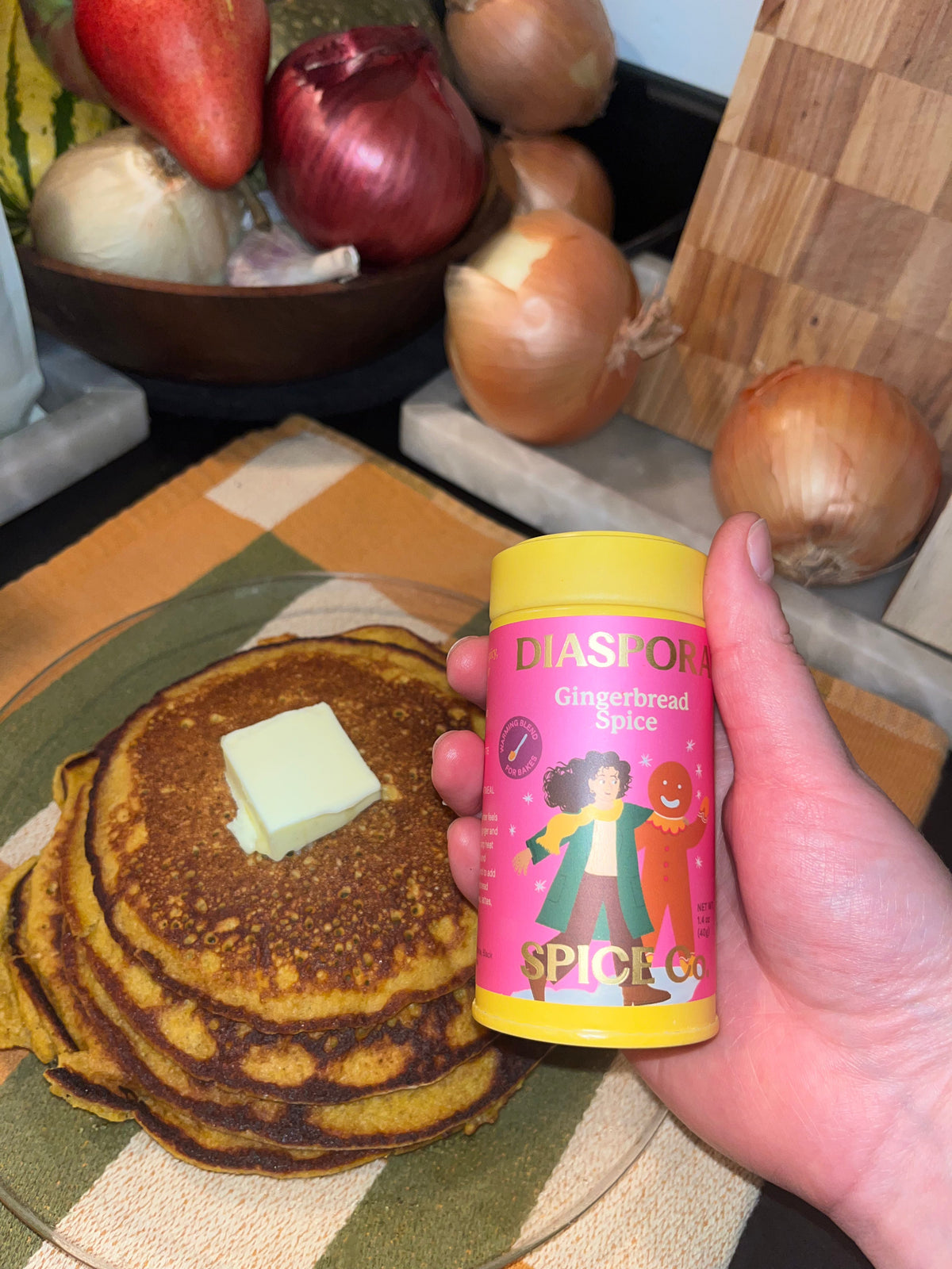 stack of pancake and tin of Diaspora Co. Gingerbread Spice in a hand with a background of fall veggies, a cutting board and a green, white and marigold tea towel 