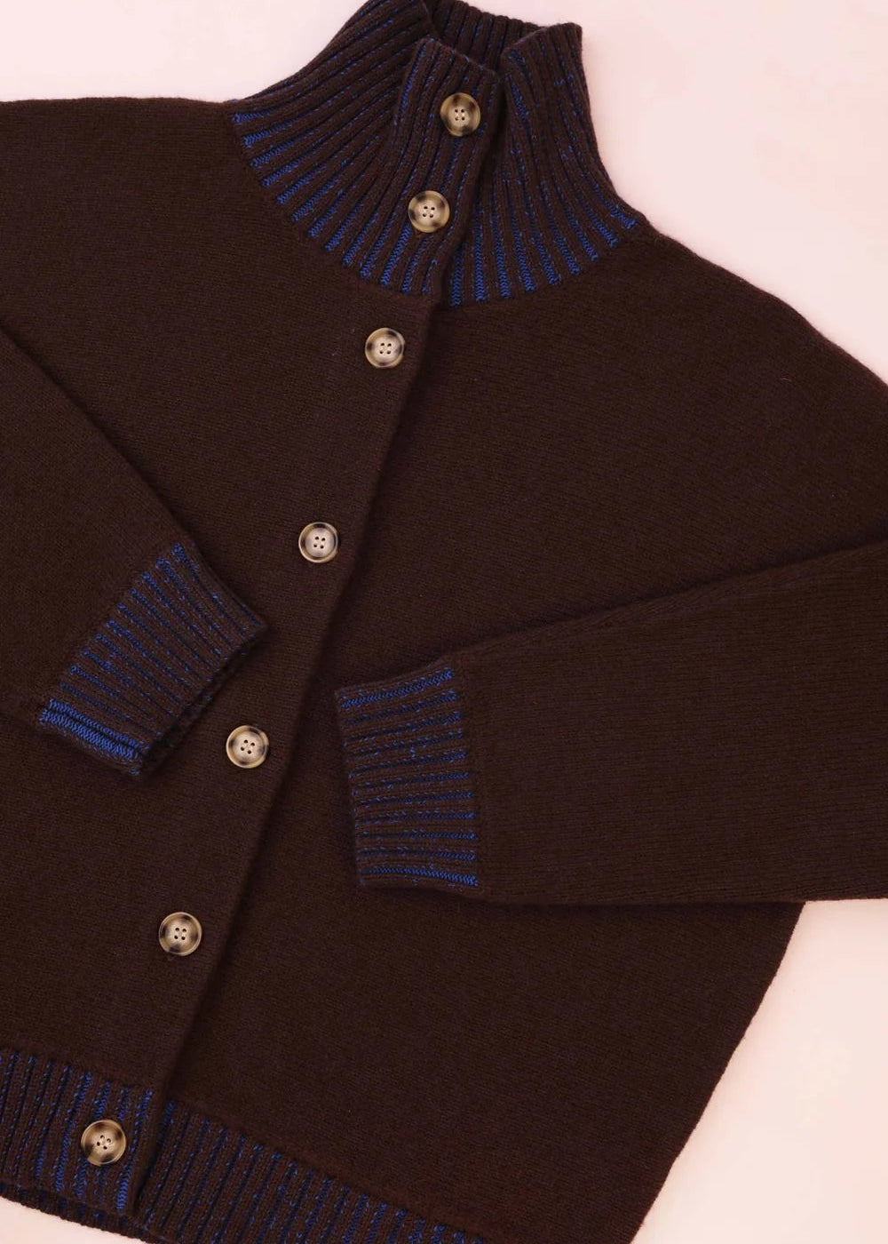 A dark brown knit cardigan sweater with a mock neck, button closure, and ribbed trim in blue and brown, laid flat on a light pink background.