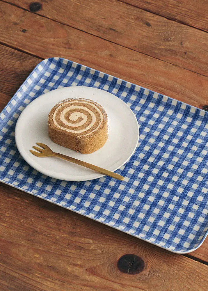 A rectangular blue and white checkered tray sits on a rustic wooden table, holding a small white plate with a slice of cake and a gold-toned fork.