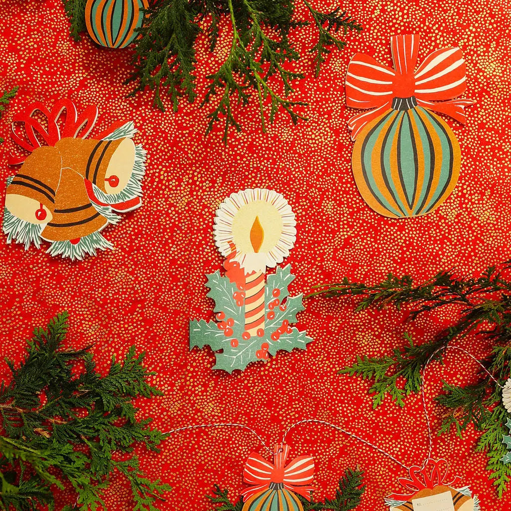 A festive flat-lay of Christmas-themed paper ornaments on a red patterned background with green pine branches. The ornaments include a striped ornament with a bow, a candle in a holly leaf, and other holiday shapes in red, orange, yellow, green, and white.