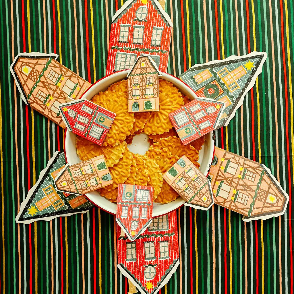 A bowl filled with golden, cookies arranged in a circular pattern, surrounded by six hand-cut gingerbread house cards with colorful windows and roofs, set against a green, red, and white striped background.