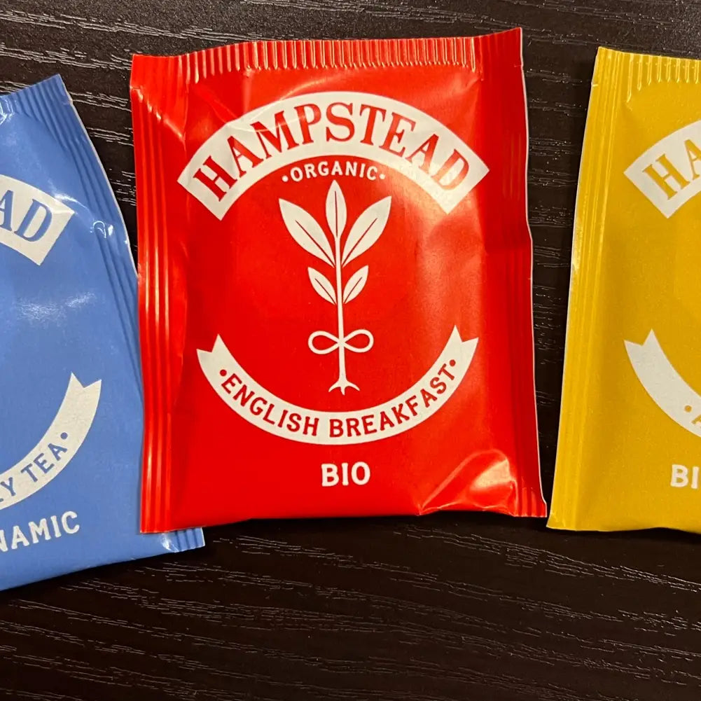 Three Hampstead Organic tea packets in red, blue, and yellow on a dark surface.