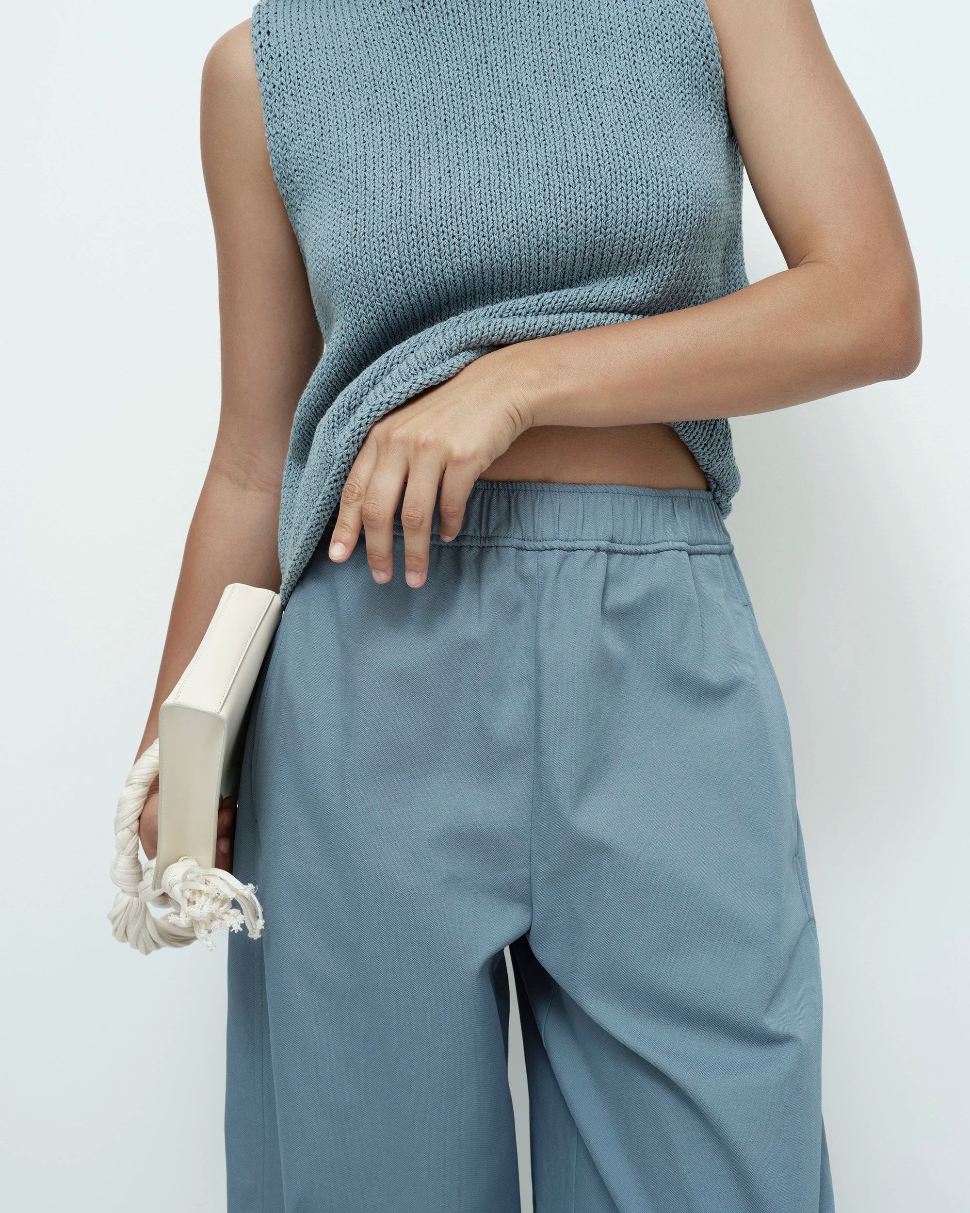 Person wearing a light blue sleeveless top and pants, holding a white clutch 