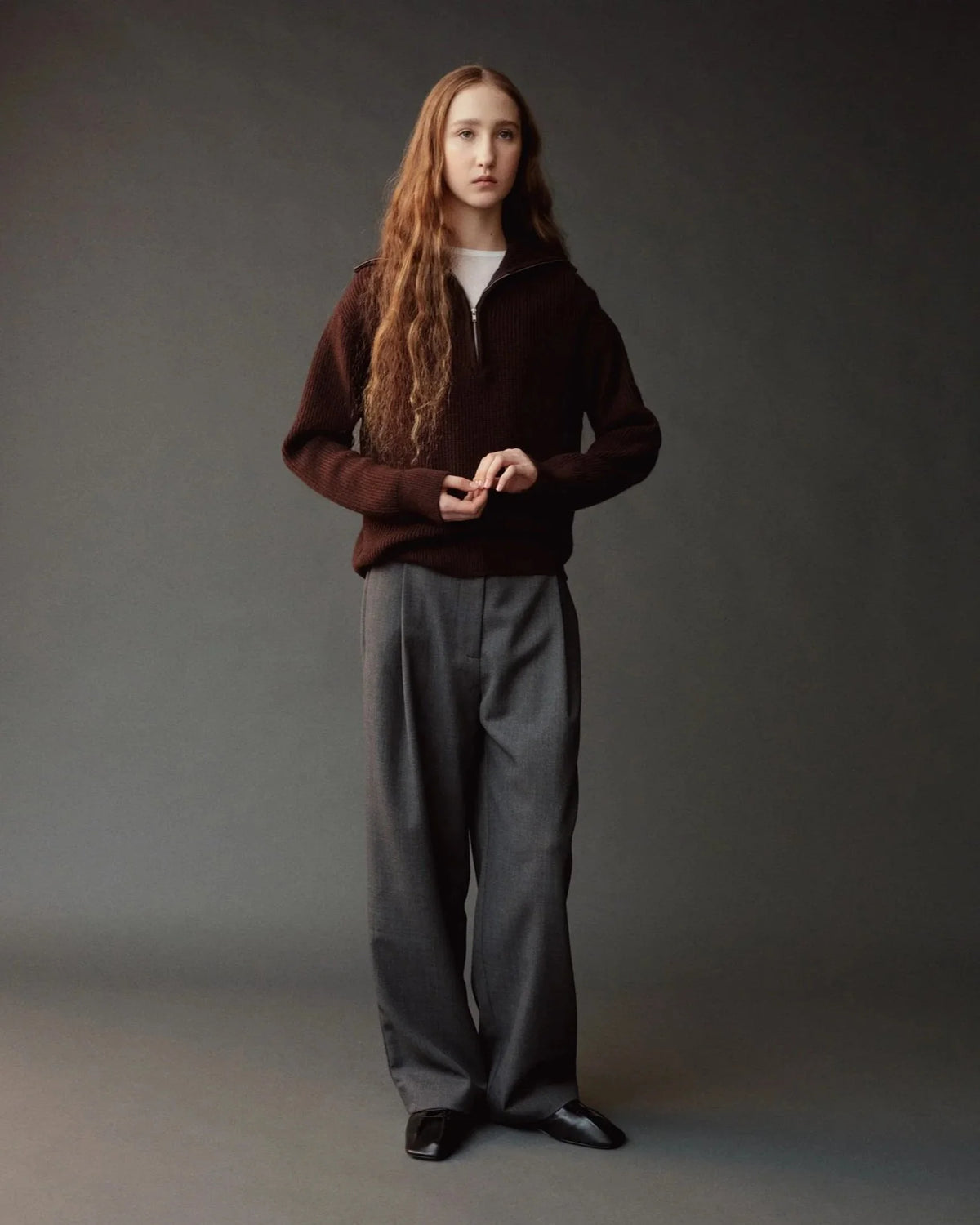 A model with long light brown hair stands against a dark gray studio background wearing a dark brown half-zip sweater, loose gray trousers, and black shoes.