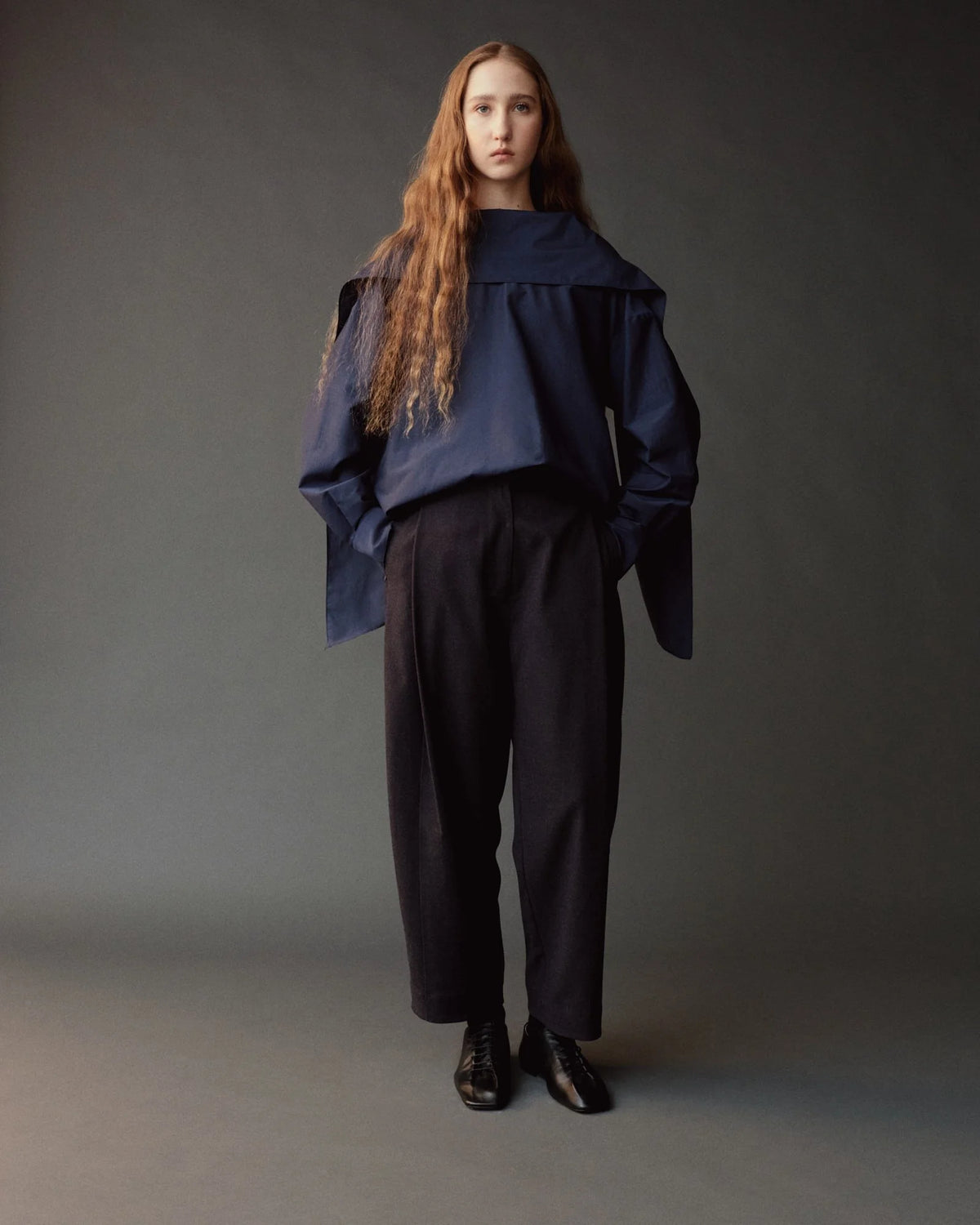 A model stands against a dark gray studio backdrop wearing a dark navy blue oversized long-sleeve top with wide layered shoulders, dark wide-leg trousers, and black shoes.
