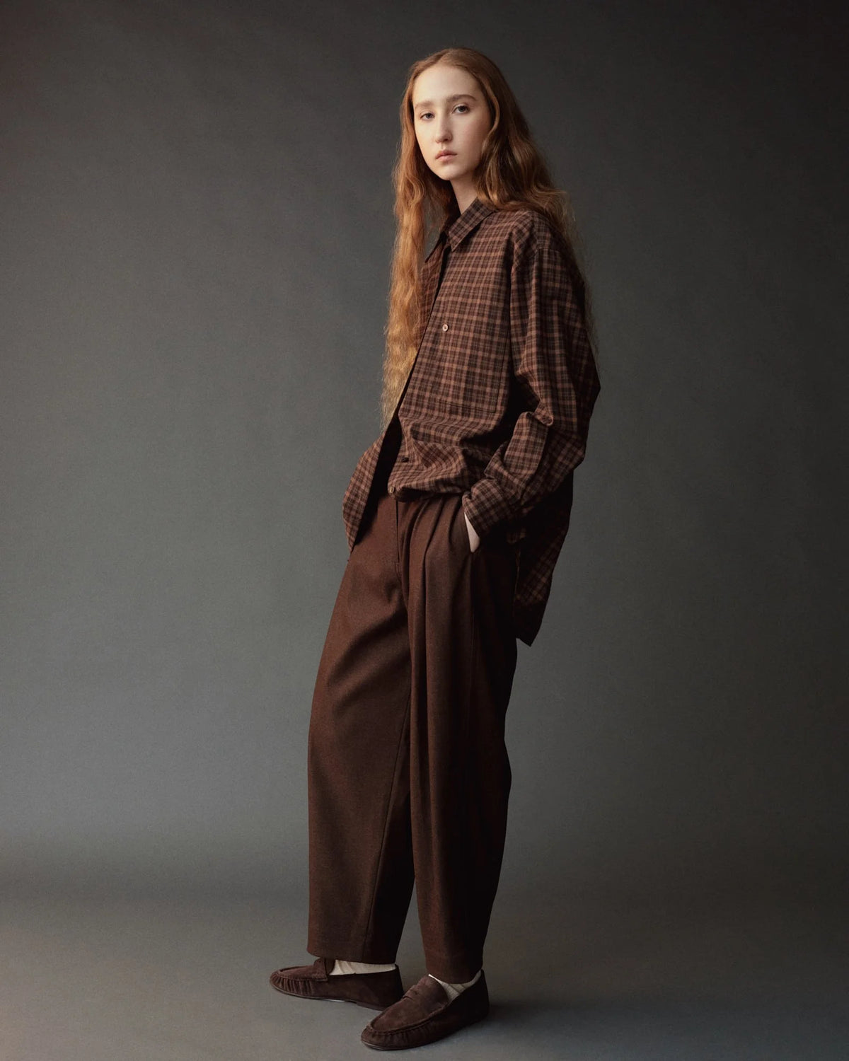 A model stands in a studio wearing a brown and dark plaid button-up shirt, matching brown wide-leg trousers, and brown slip-on shoes against a dark gray background.