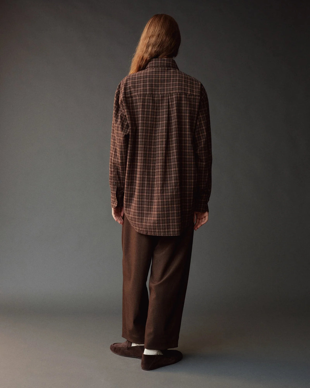 A person shown from the back wearing a brown plaid button-up shirt and matching brown trousers, standing against a dark gray studio background.