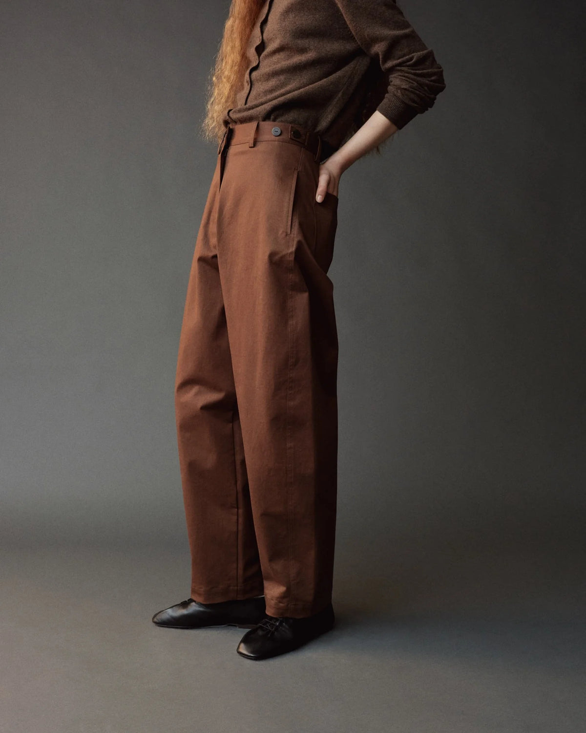 A model wearing a pair of wide-leg brown trousers, styled with a dark brown long-sleeve shirt and black leather shoes, standing against a dark gray studio background.