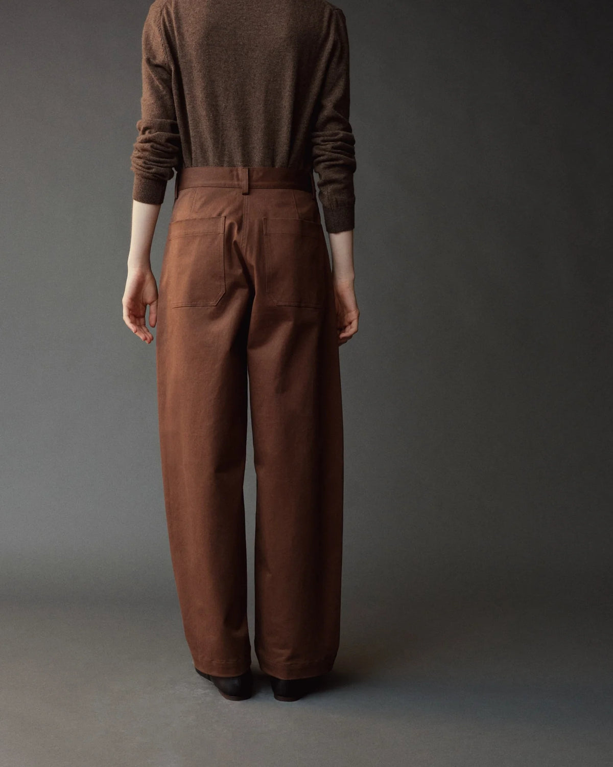 A person shown from the back wearing brown wide-leg trousers and a matching brown long-sleeve top against a dark gray studio background.