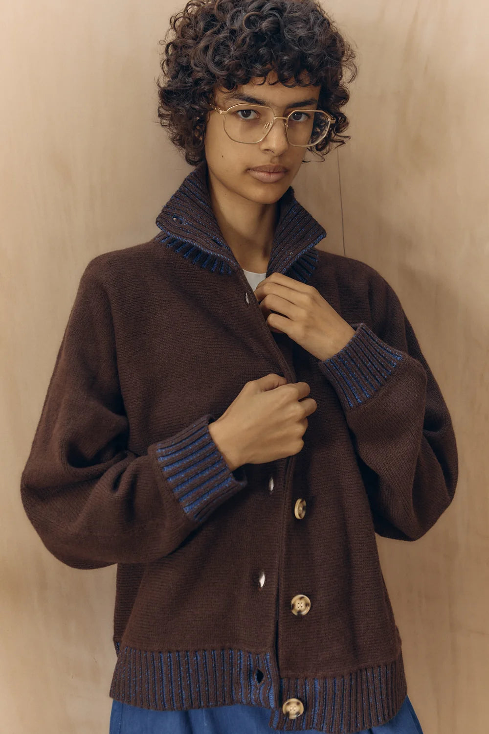 A model wearing a dark brown knit cardigan with blue ribbed trim and gold buttons, posed against a light wood-paneled background.