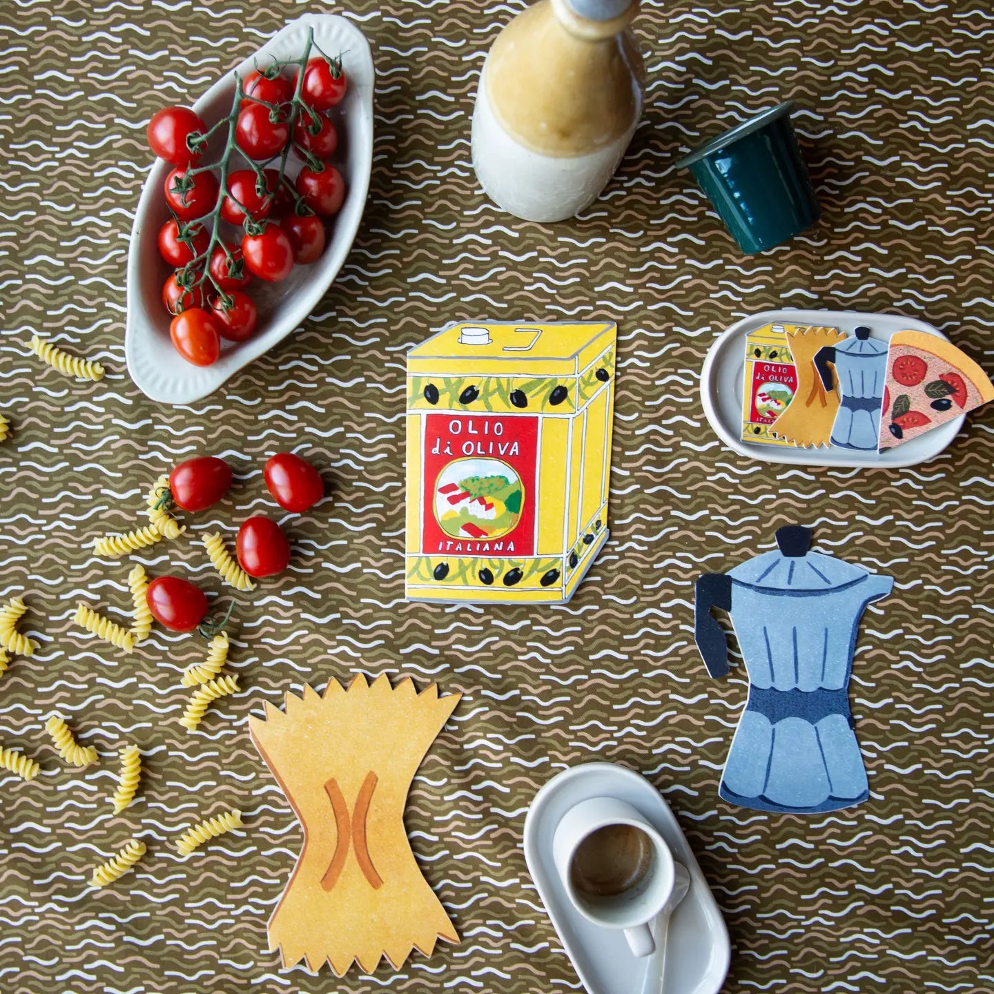 A styled flat-lay of kitchen-themed paper cutouts and props on a brown wavy-patterned surface, including a yellow olive oil box labeled 'Olio di Oliva Firenze,' a blue moka pot silhouette, a yellow pasta cutter, cherry tomatoes, pasta, a bottle, and a coffee cup.