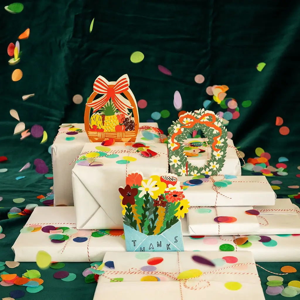 Three wrapped gift boxes decorated with colorful paper pop-art toppers, including a basket with fruit, a floral wreath, and a flowers card reading THANKS, arranged on a dark green fabric backdrop with scattered confetti.