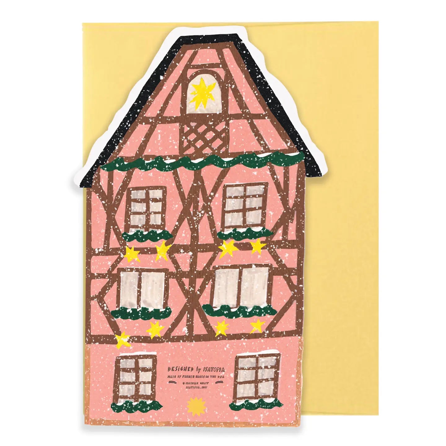 A decorative house-shaped card or sticker featuring a pink half-timbered house with brown beams, green garlands, yellow stars, and a white snowy outline, placed in front of a pale yellow background.