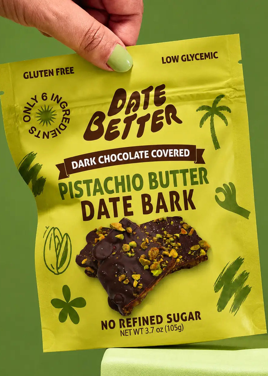 A hand with light green nail polish holds a bright yellow bag of Date Better Pistachio Butter Date Bark against a solid green background. The packaging features dark brown text and illustrations, and a photo of the chocolate-covered date bark on the front.