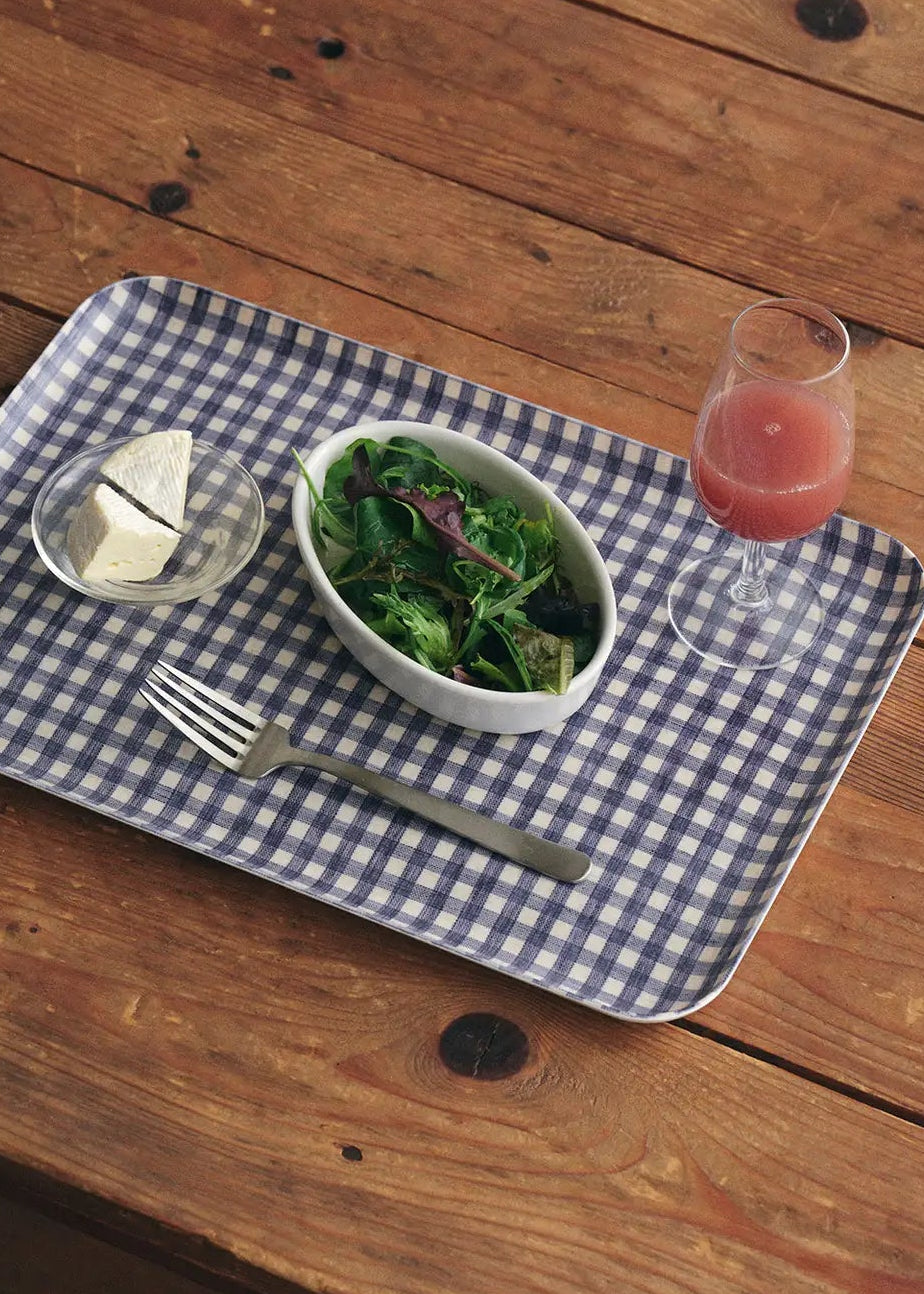 Checkered blue and white tray with a bowl of salad, glass of juice, and fork on a wooden table.