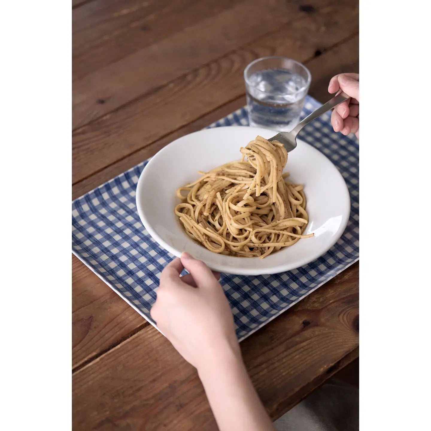 A white bowl of spaghetti pasta on a blue and white checkered placemat on a wooden table, with a fork lifting the pasta and a glass of water in the background.
