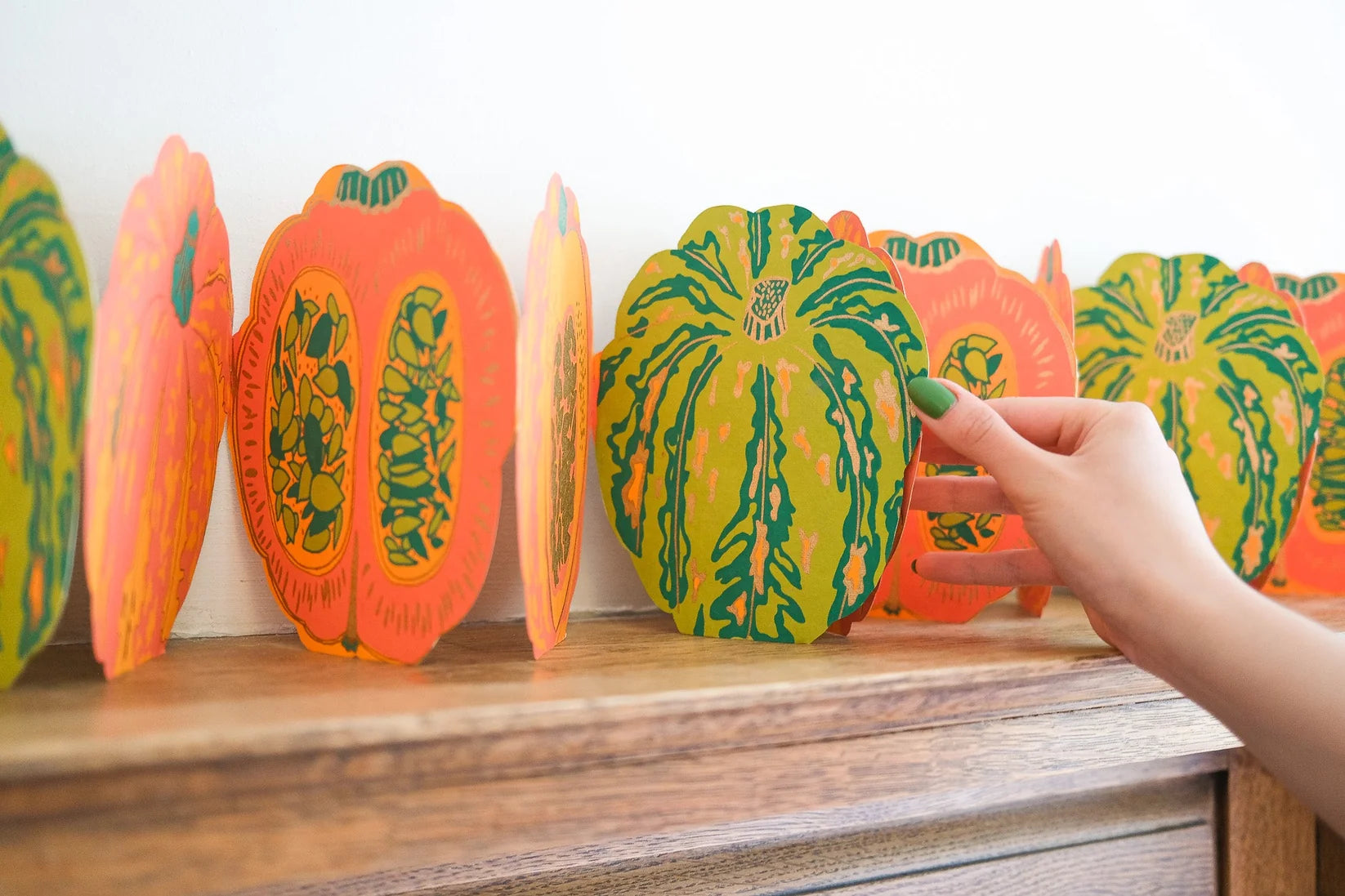 A hand with green nail polish gently touches a colorful paper pumpkin decoration on a wooden shelf, with other matching paper pumpkins lined up against a plain white wall.