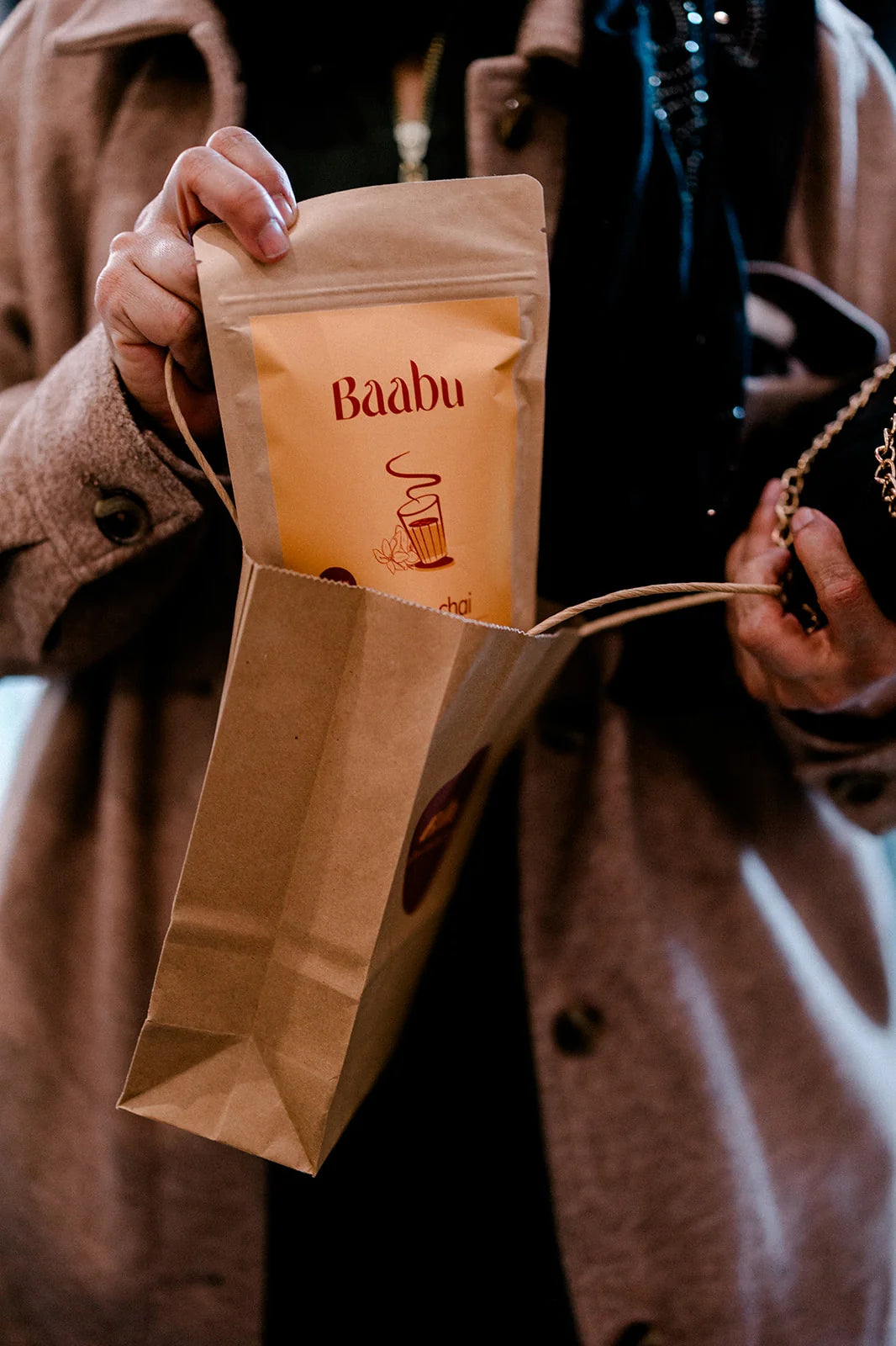 A person in a brown coat holds a kraft paper bag containing a smaller pouch labeled 'Baabu' in warm orange-brown tones, photographed in a real-world setting.