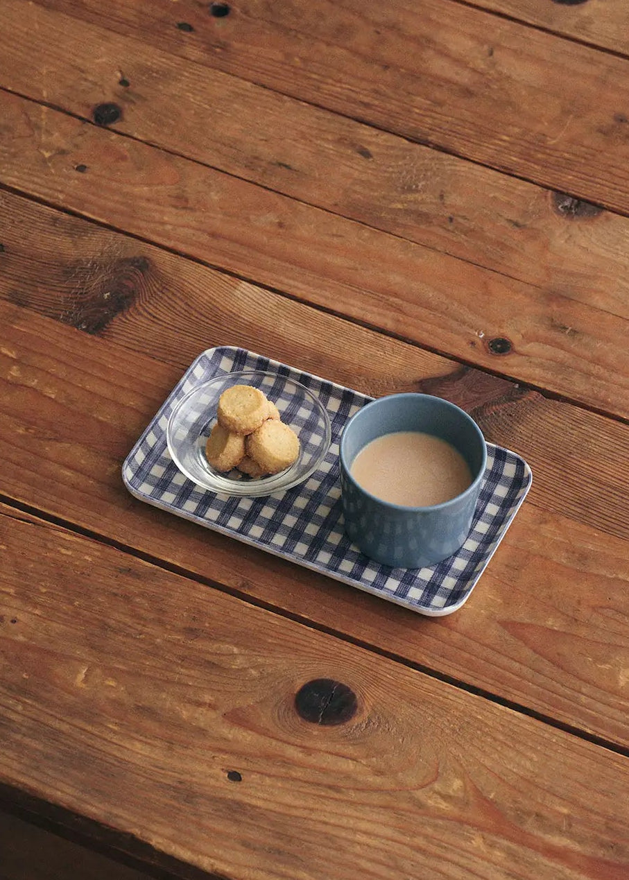 A rectangular tray with a blue and white gingham pattern holds a small glass bowl of cookies and a blue mug filled with a light brown beverage, set on a rustic wooden table.