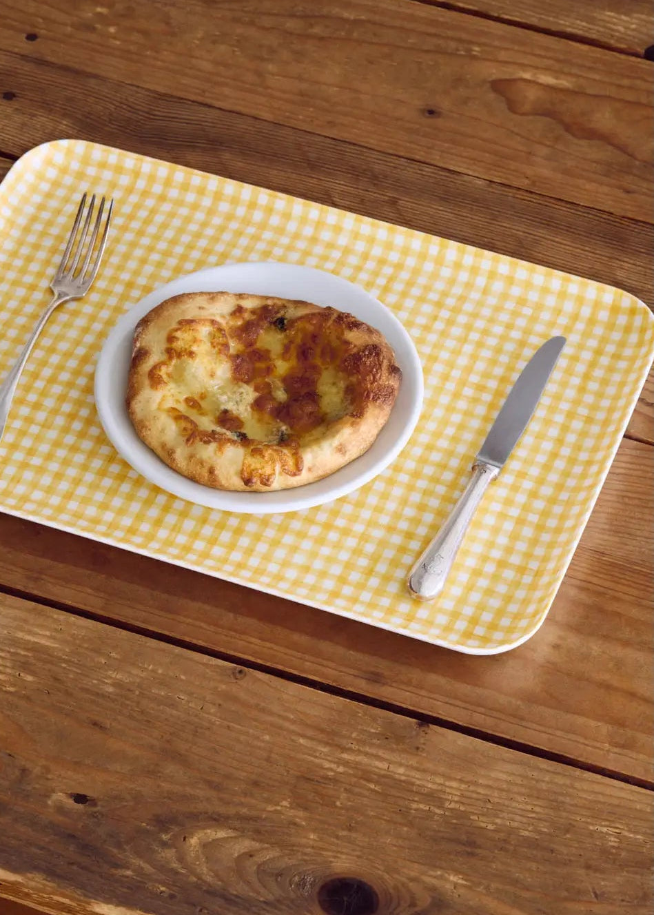 Small pizza on a white plate with a fork and knife on a yellow checkered placemat over a wooden table.