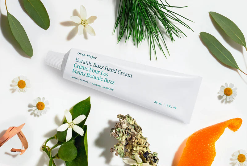 Ursa Major Botanic Buzz Hand Cream surrounded by greenery and flowers on a white background