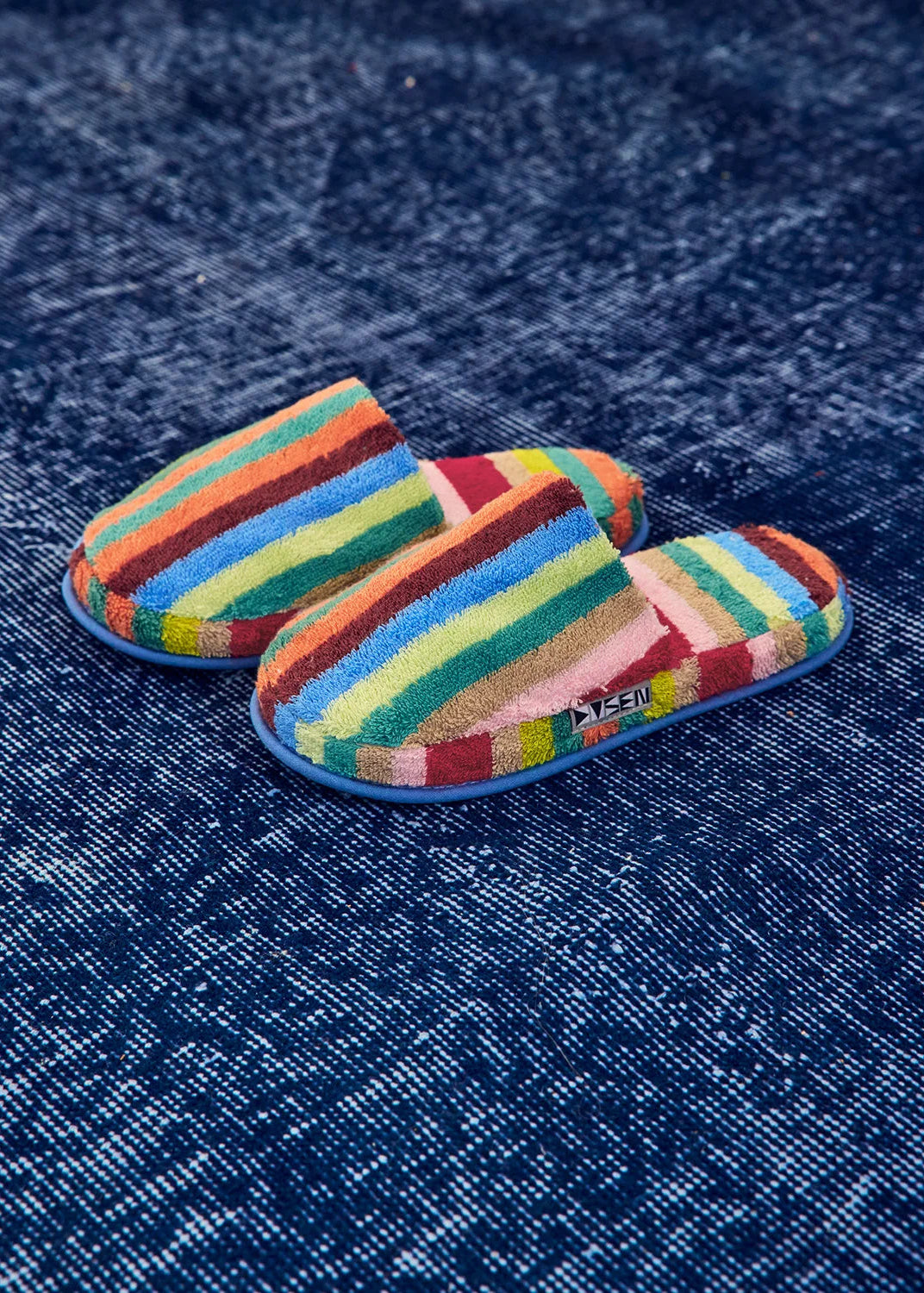 A pair of colorful striped slip-on slippers with a blue sole, placed on a textured dark blue fabric background. The slippers feature multiple horizontal stripes in red, orange, yellow, green, blue, and pink, with a small white logo on the side.