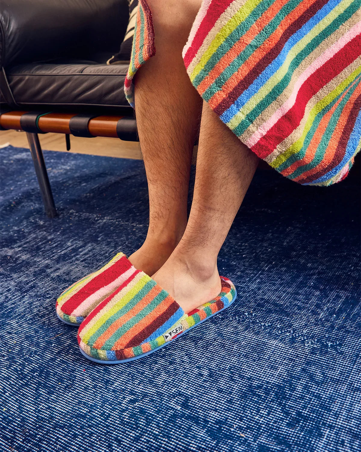 A person sitting on a black leather sofa wearing colorful striped bathrobes and matching striped slip-on slippers on a textured blue rug.