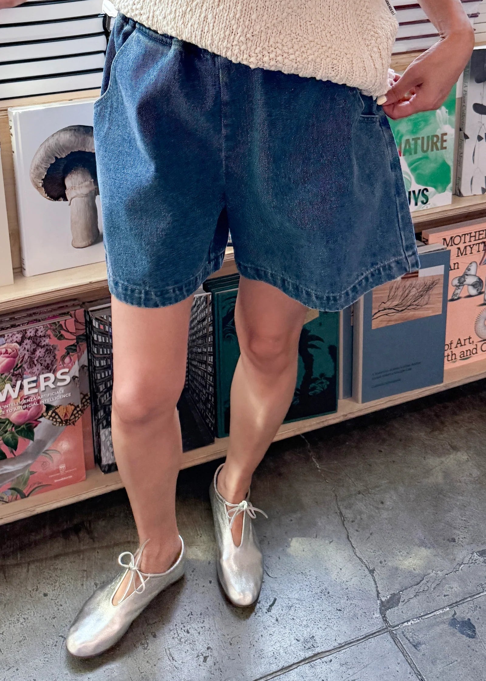 Person wearing blue shorts and silver shoes standing in front of a bookshelf.
