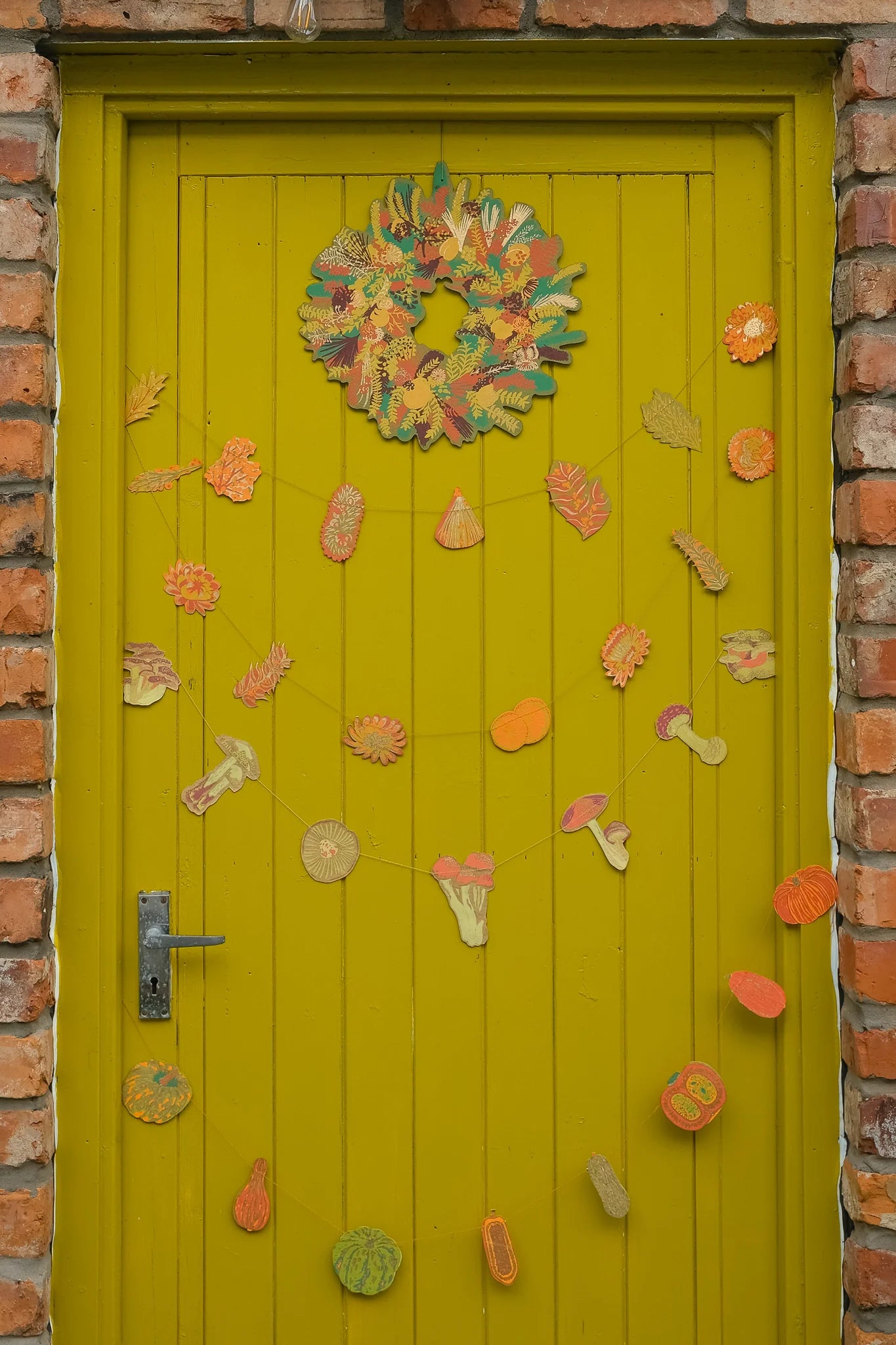 A bright yellow-green wooden door decorated with autumn-themed paper crafts, including a colorful wreath and string garlands of leaves, acorns, and mushrooms, set against a red brick wall.