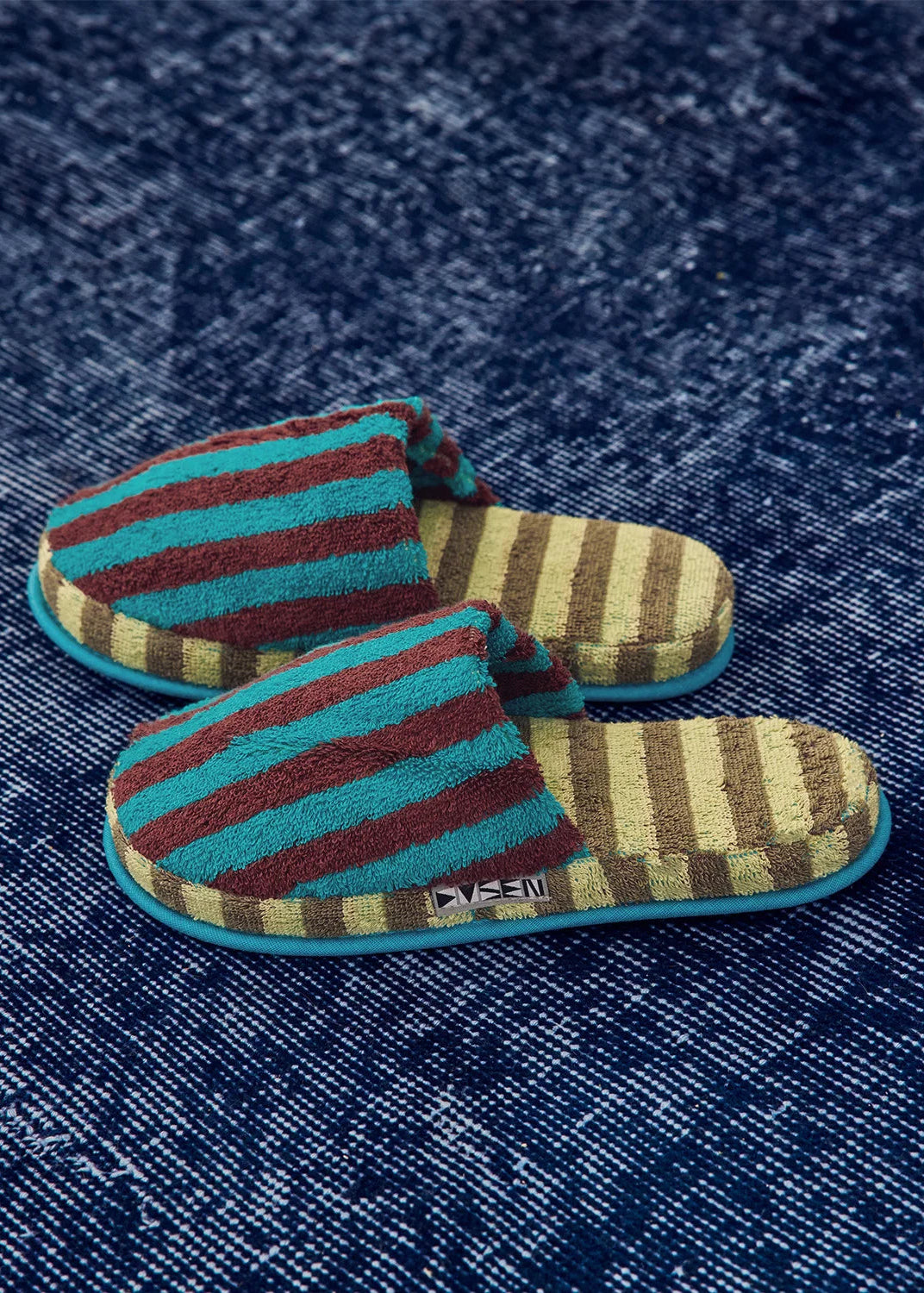 A pair of striped slip-on slippers with a plush texture, featuring teal, burgundy, and beige horizontal stripes on the upper, and beige and brown stripes on the footbed, resting on a dark blue textured fabric background.