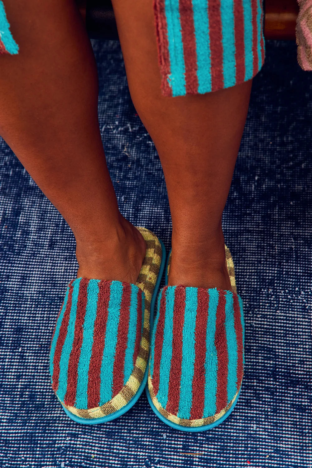 A close-up of a person wearing striped slip-on slippers with turquoise, rust red, and gold vertical stripes, standing on a blue textured rug.