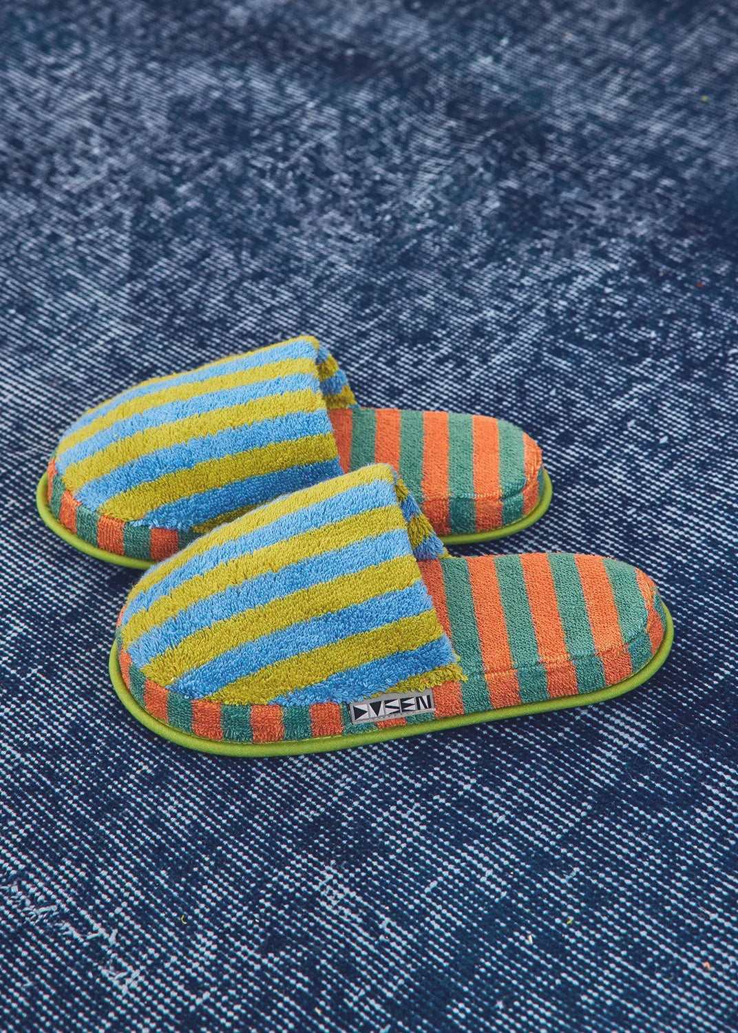 A pair of striped slip-on slippers with blue, yellow, orange, and green horizontal stripes, displayed on a textured blue denim background.