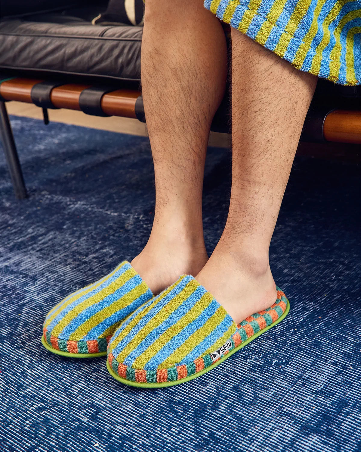 A person sitting on a bench wearing colorful striped terry cloth slip-on slippers with blue, yellow, orange, and green stripes, standing on a textured blue rug.