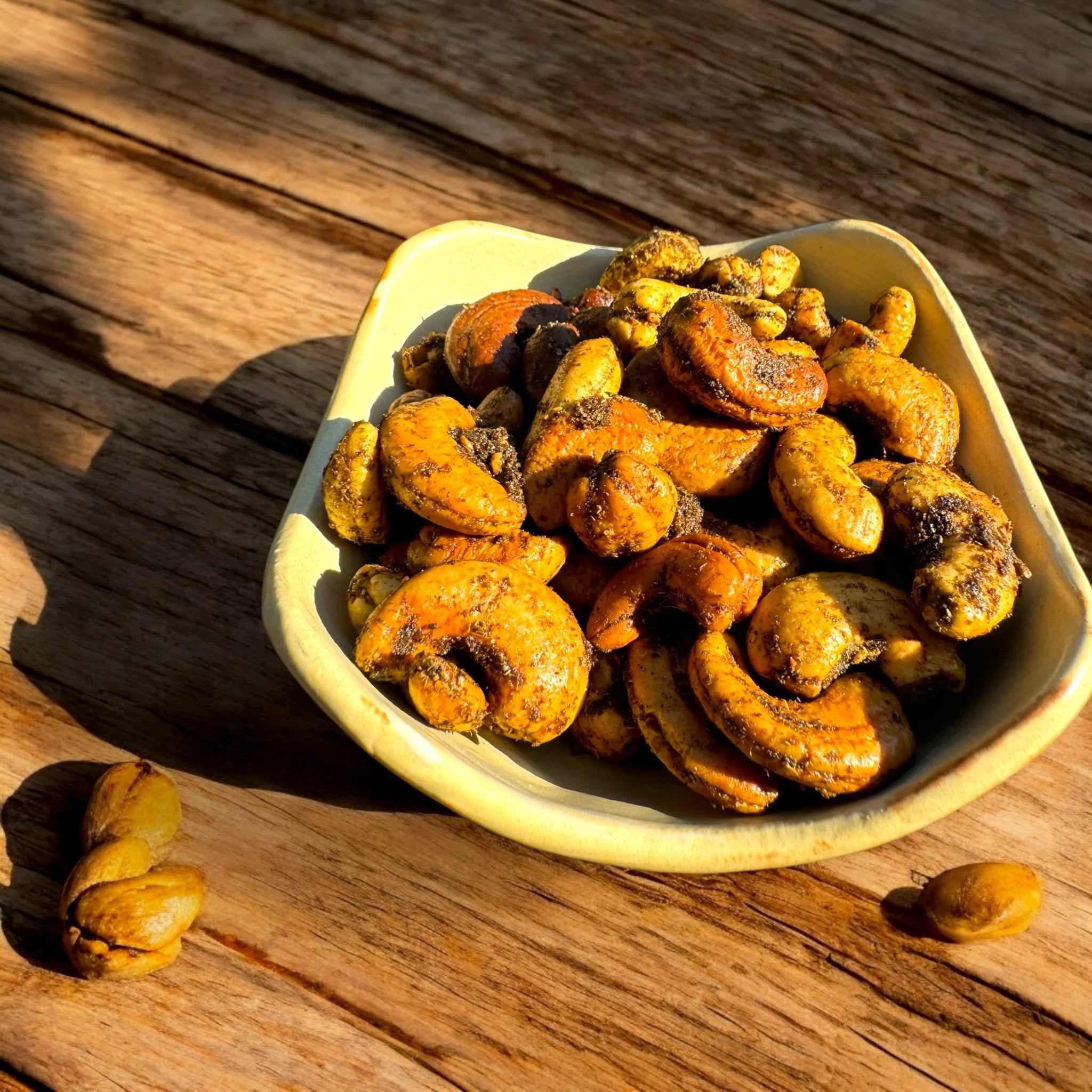 Roasted cashews in a small light beige ceramic bowl, placed on a rustic wooden surface with a few whole cashews scattered nearby under warm sunlight.