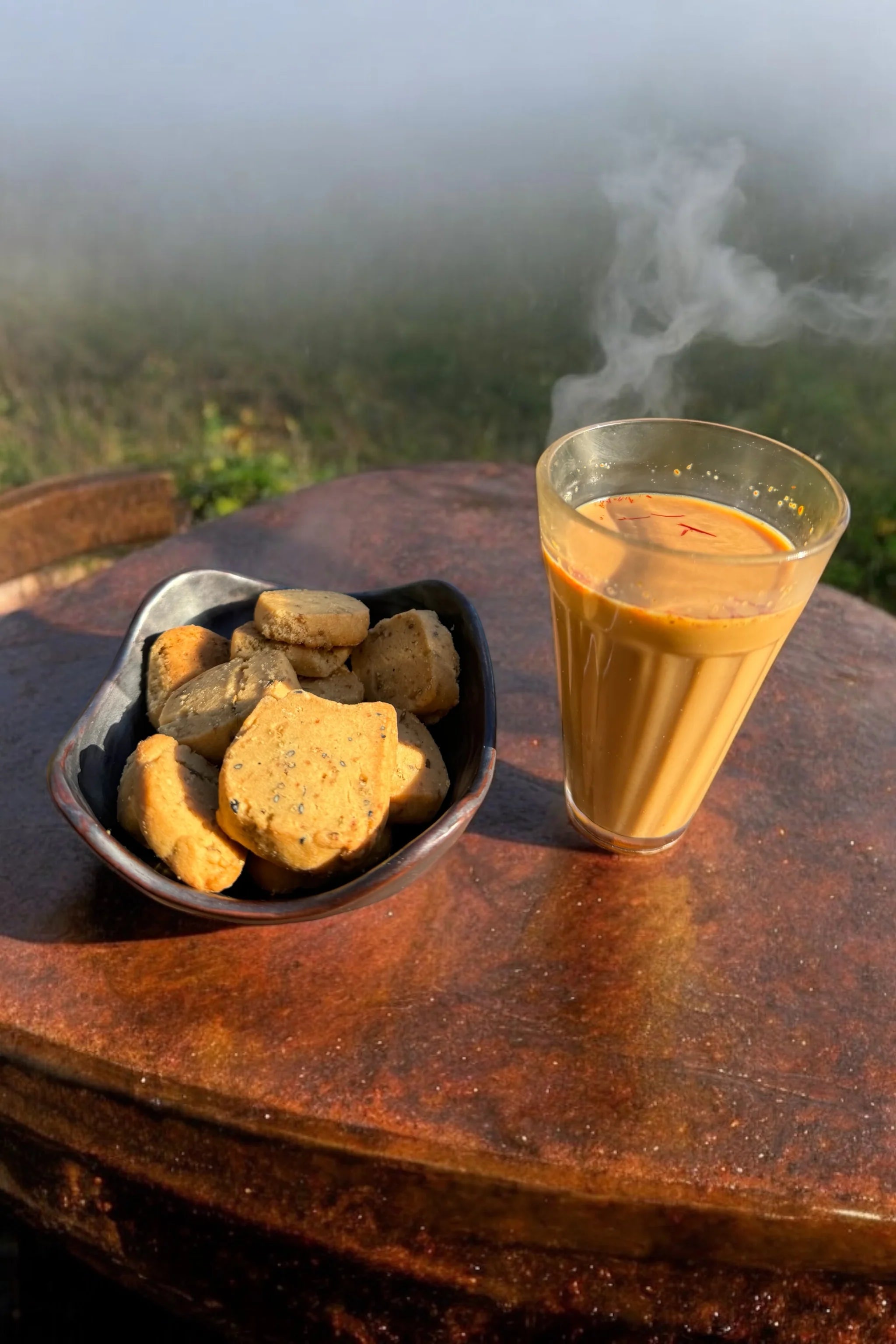 A glass of steaming orange-colored beverage and a small bowl of cookies on a rustic wooden table, with a misty outdoor landscape in the background.