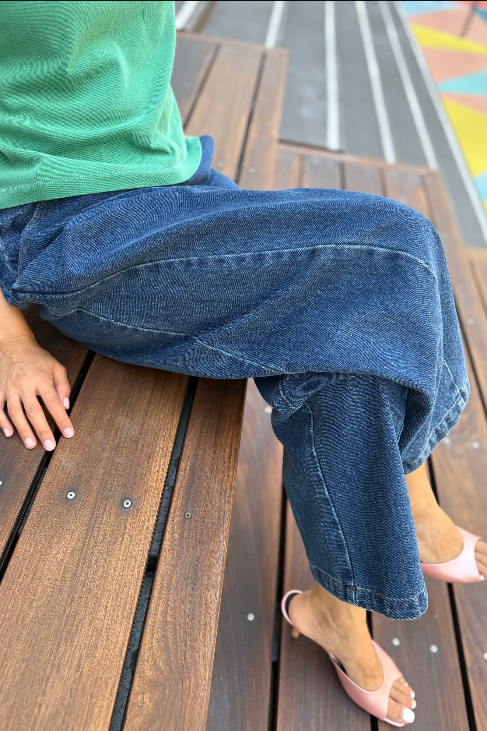 Person wearing blue jeans sitting on a wooden bench with a colorful playground in the background.