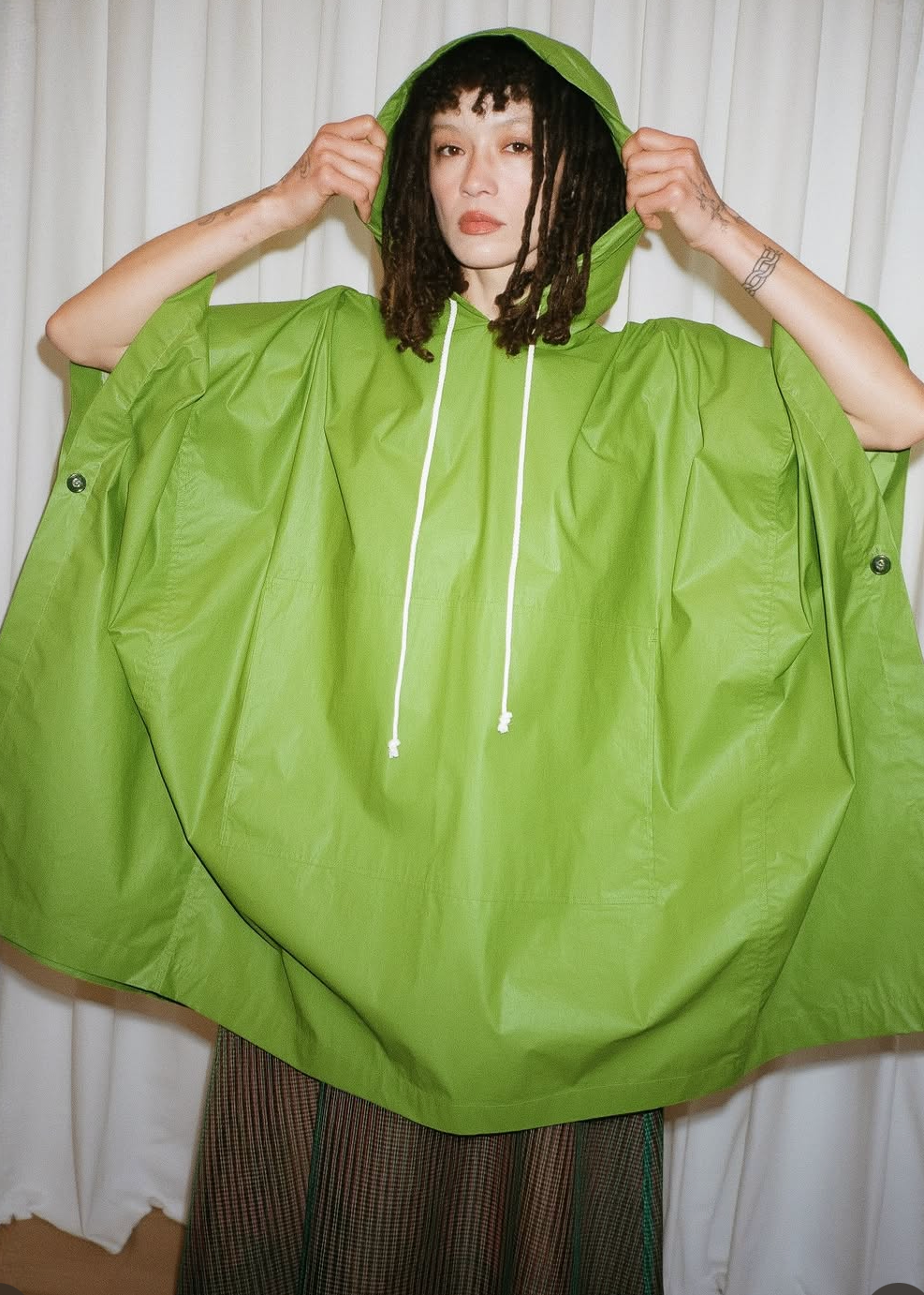 A person wearing a bright lime green hooded rain poncho with white drawstrings, standing indoors against a light curtain background. The photo appears to be a social media post screenshot with a 'View shop' banner at the bottom.