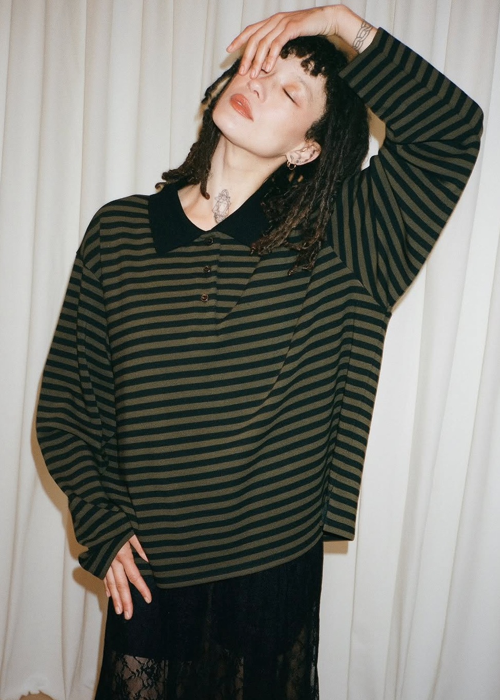A woman modeling a dark green and black horizontally striped long-sleeve knit top with a black collar, set against a white curtain background. The image is a screenshot from a social media post.