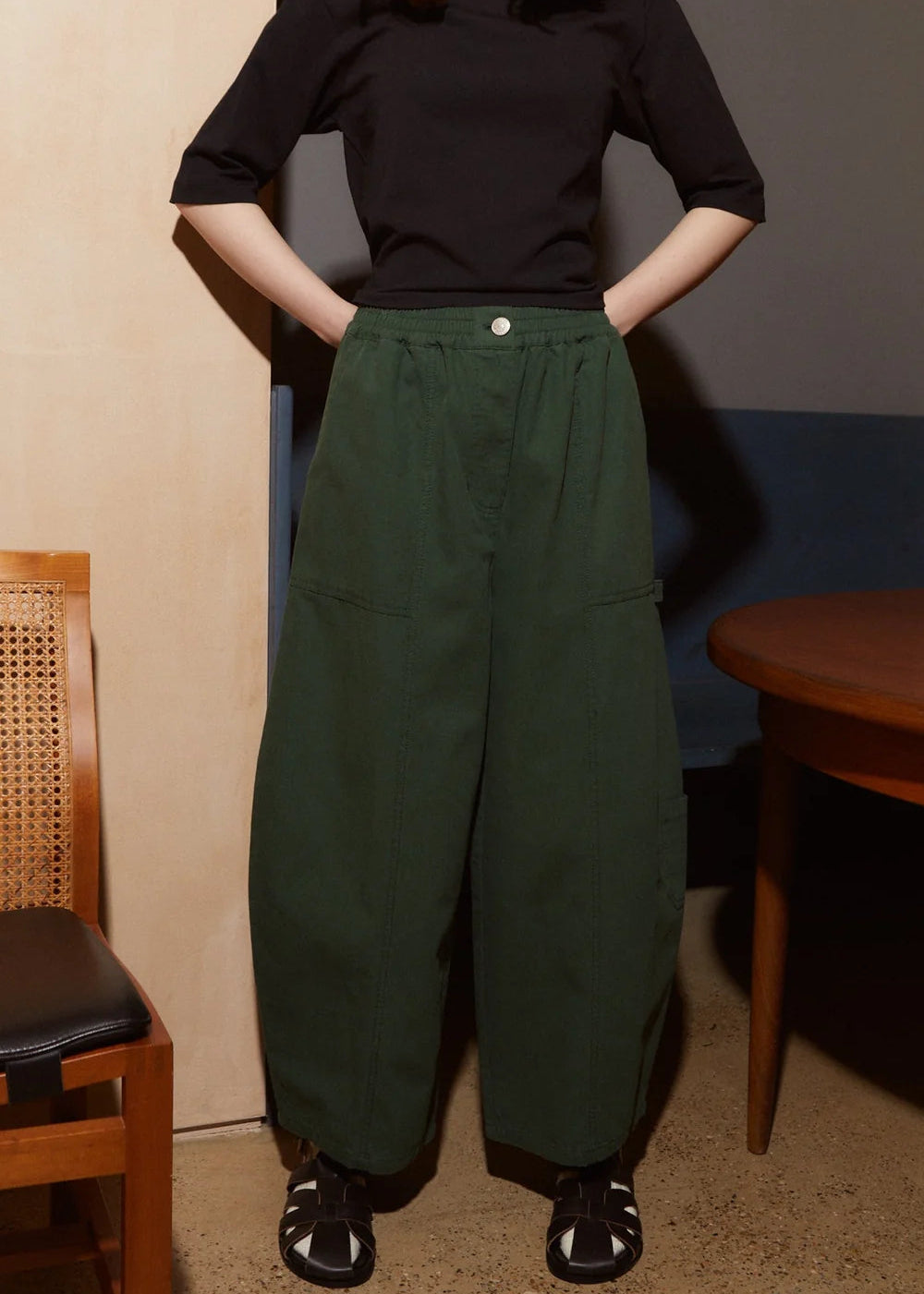 A person wearing dark green wide-leg cargo pants with a black short-sleeve top and black sandals, standing indoors with hands in pockets.