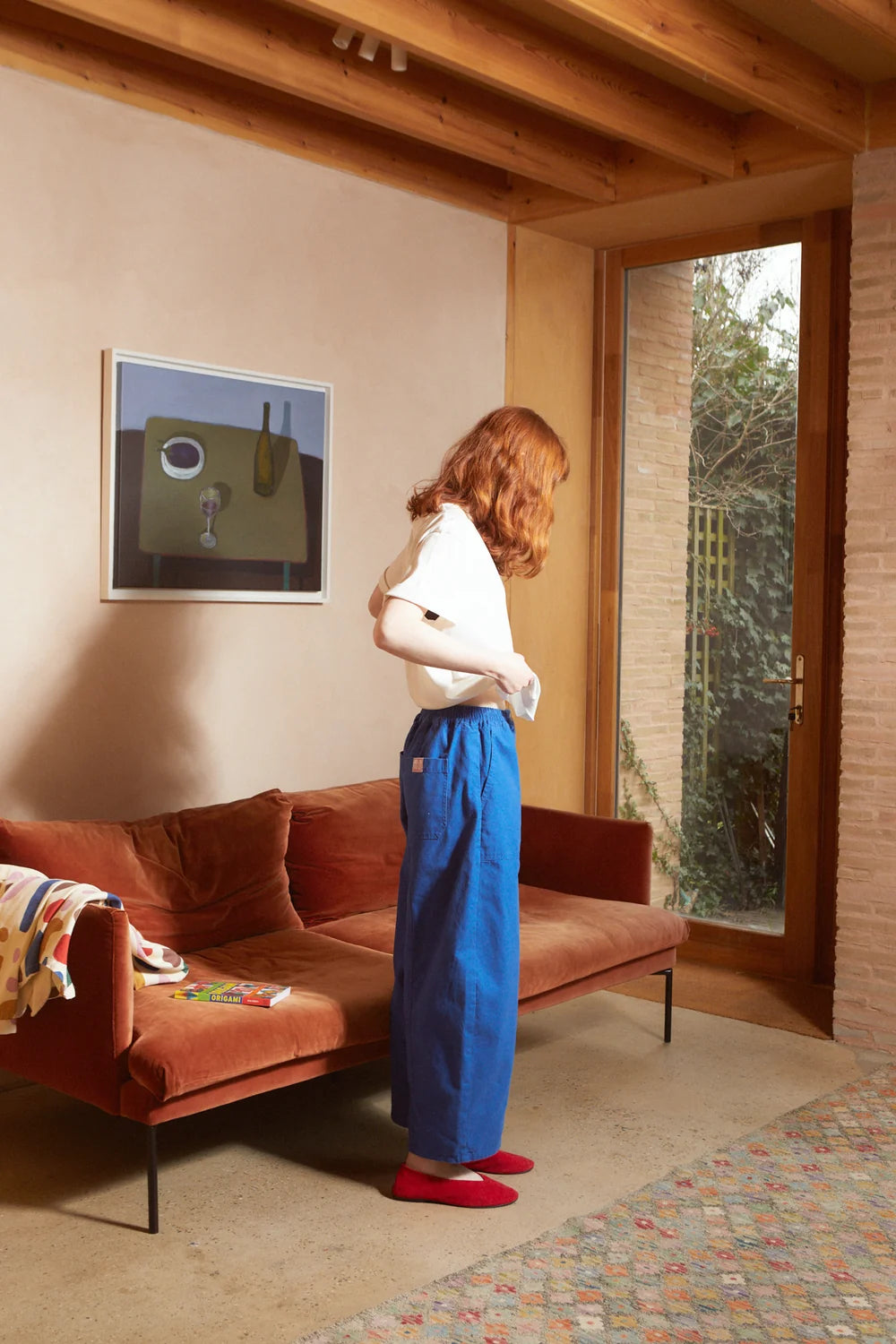 A woman with red hair stands in profile in a living room, wearing a white short-sleeve top, high-waisted wide-leg blue trousers, and red shoes. The room features a terracotta-colored sofa, a framed abstract painting, and a large window looking out onto greenery.