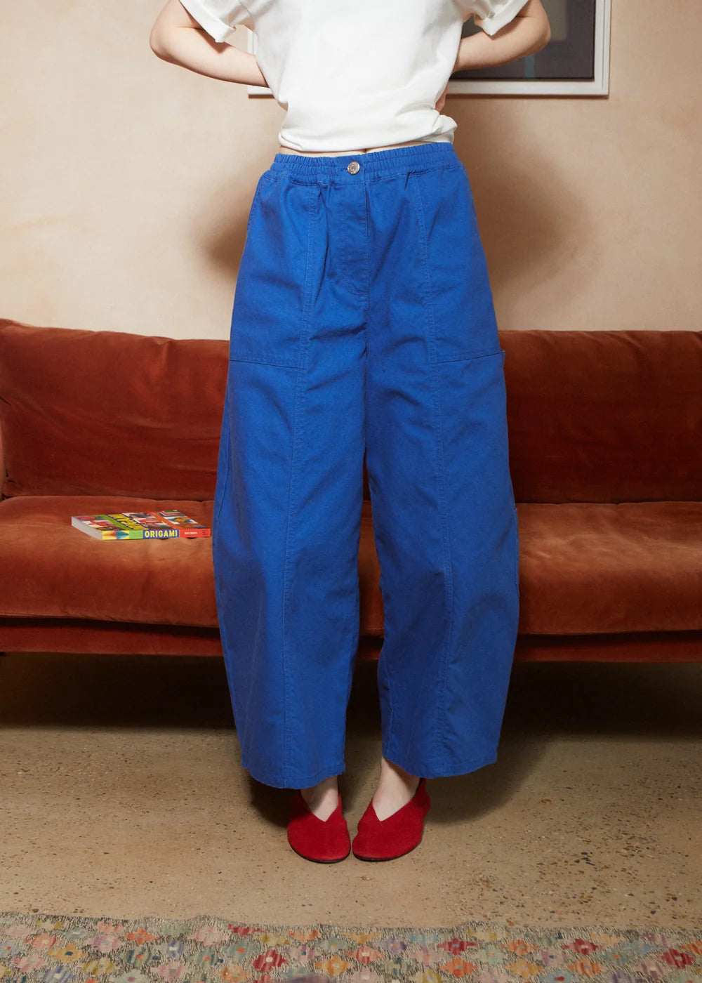 A person wearing bright royal blue wide-leg cropped pants, a white top, and red shoes, standing in front of a brown velvet sofa in an indoor room.