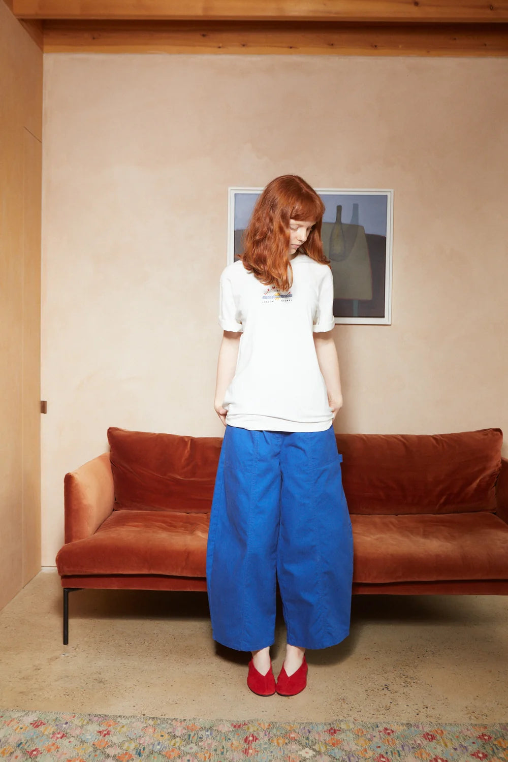 A woman with red hair stands in a room with a terracotta-colored velvet sofa, wearing a white t-shirt, wide-leg blue pants, and red shoes.