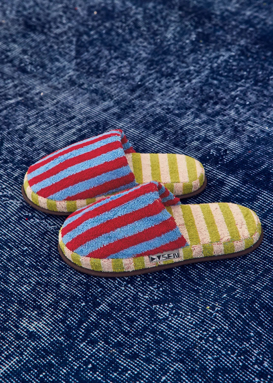A pair of slide slippers with colorful horizontal stripes in red, light blue, olive green, and cream, featuring a small black brand tag that reads 'Dusen'. They are photographed on a textured dark blue denim background.