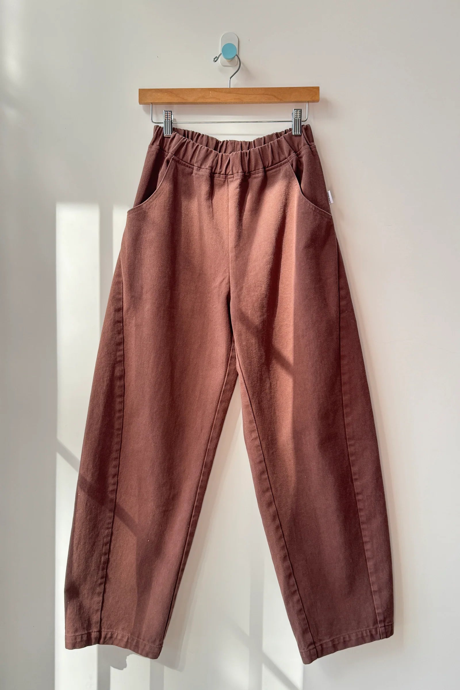 Brown pants hanging on a wooden hanger against a white wall.