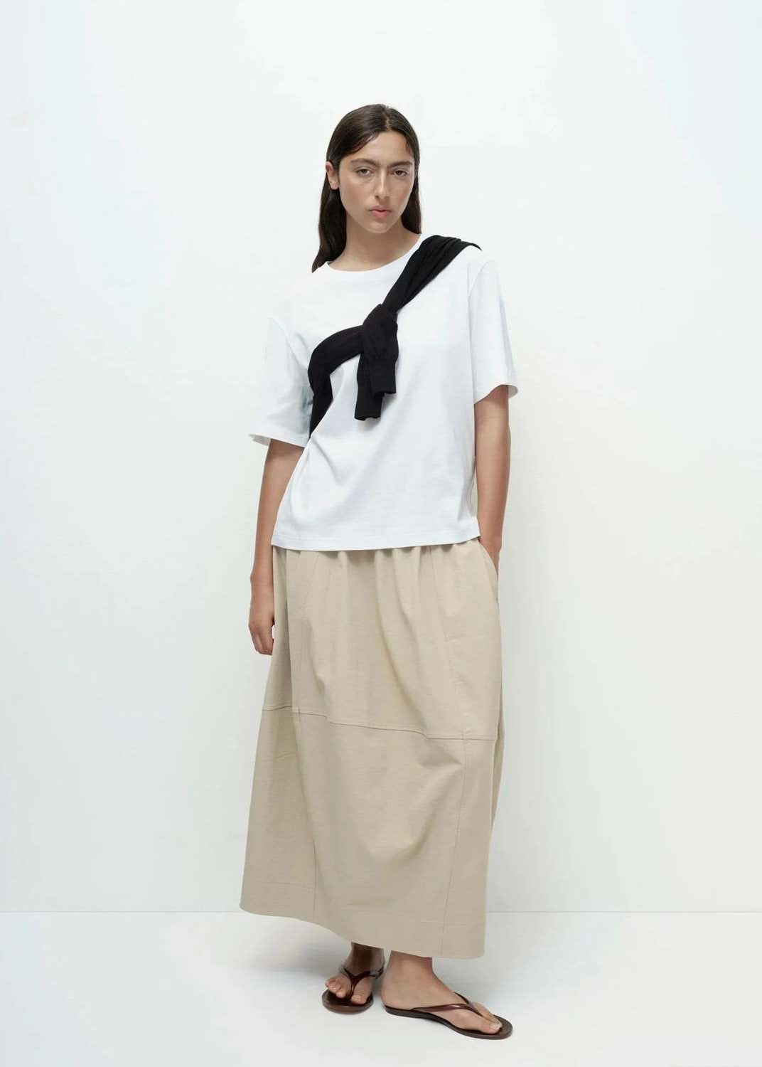 Woman wearing a white top and beige pants with a black strap over her shoulder on a white background