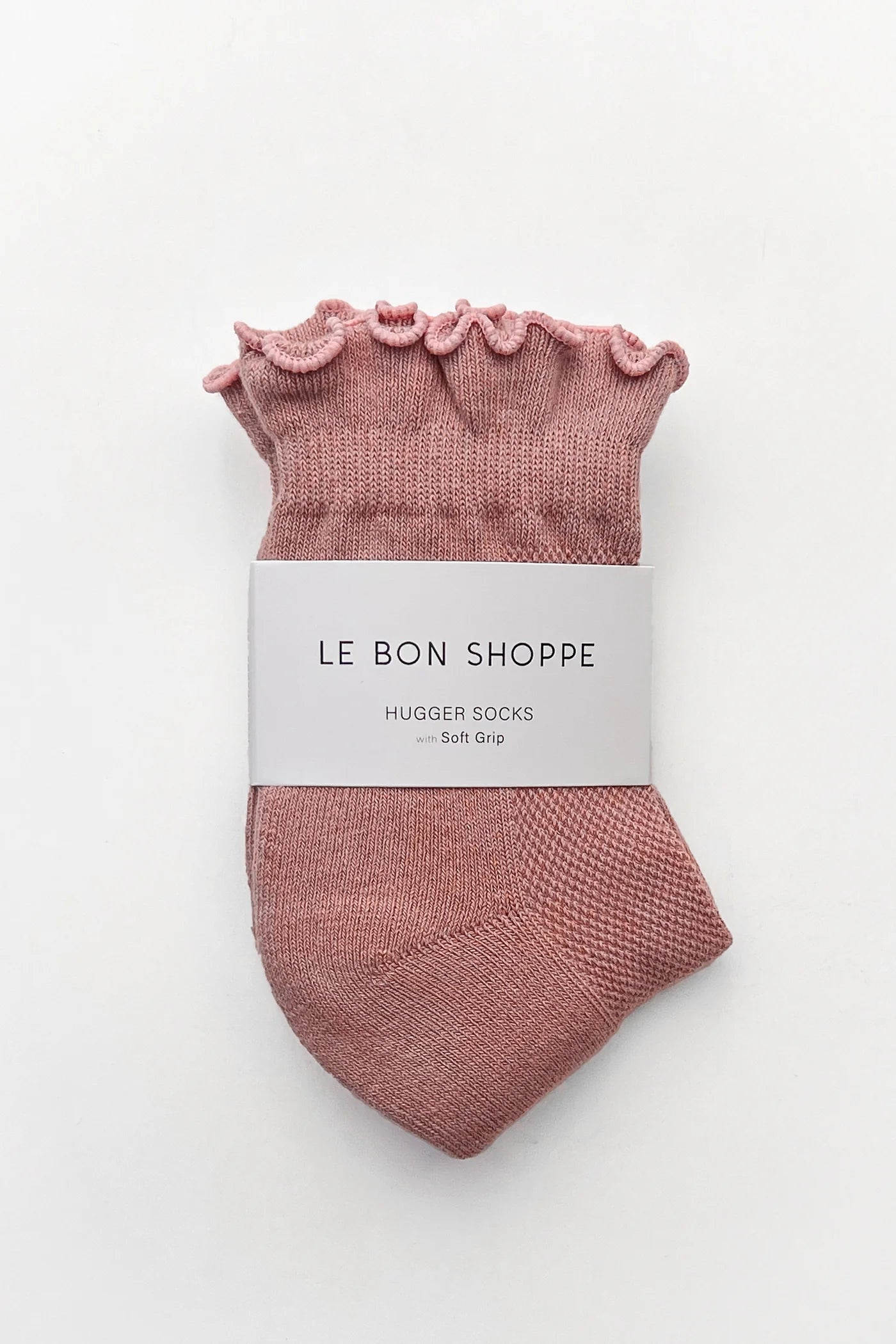A pair of dusty rose pink knit hugging socks with a ruffled top edge, wrapped in a white paper band labeled 'LE BON SHOPPE HUGGER SOCKS - Soft Grip', on a clean white background.
