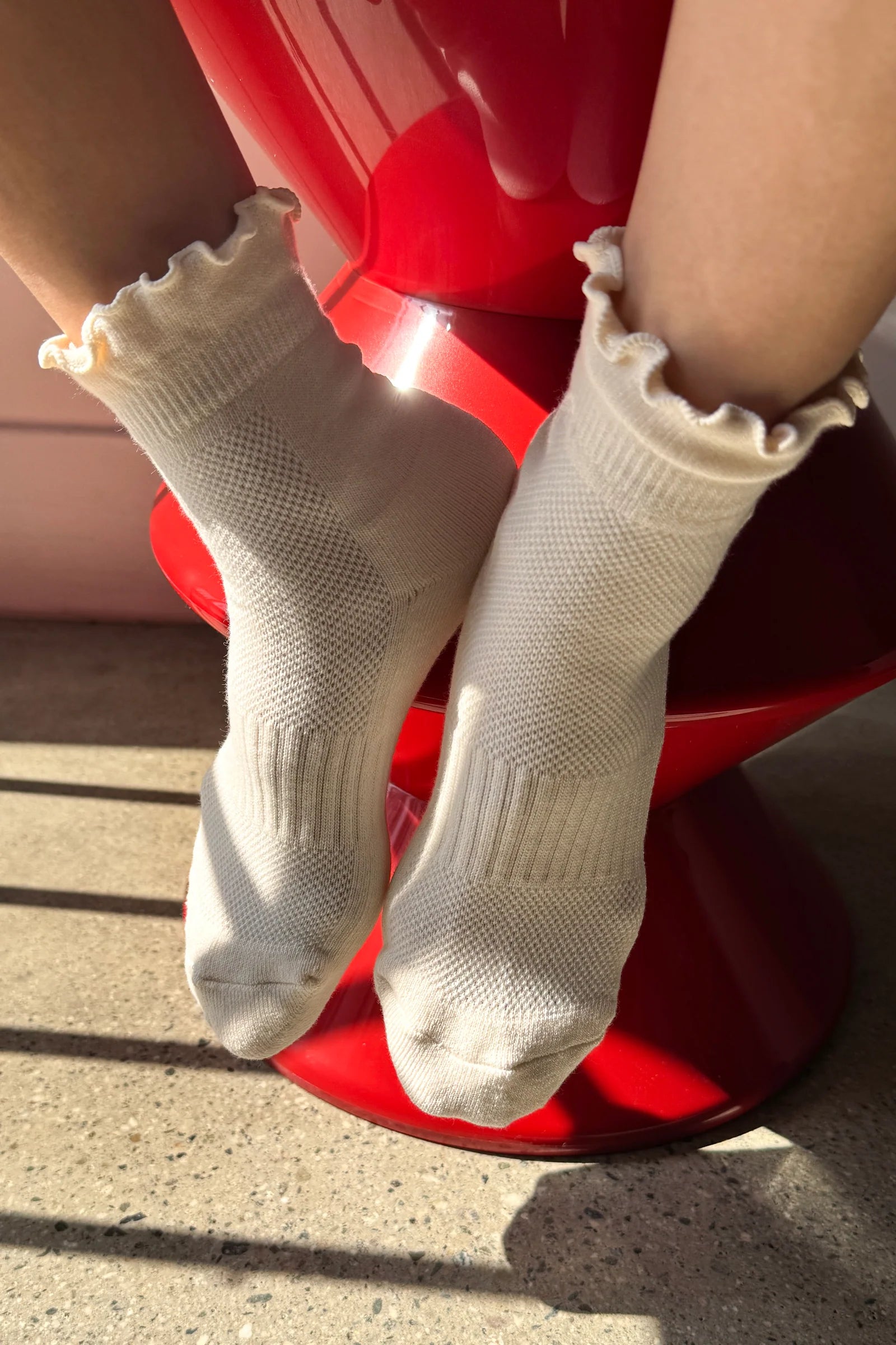 A close-up of a person's feet wearing off-white ribbed knit socks with ruffled cuffs, sitting on a glossy red chair in bright sunlight.