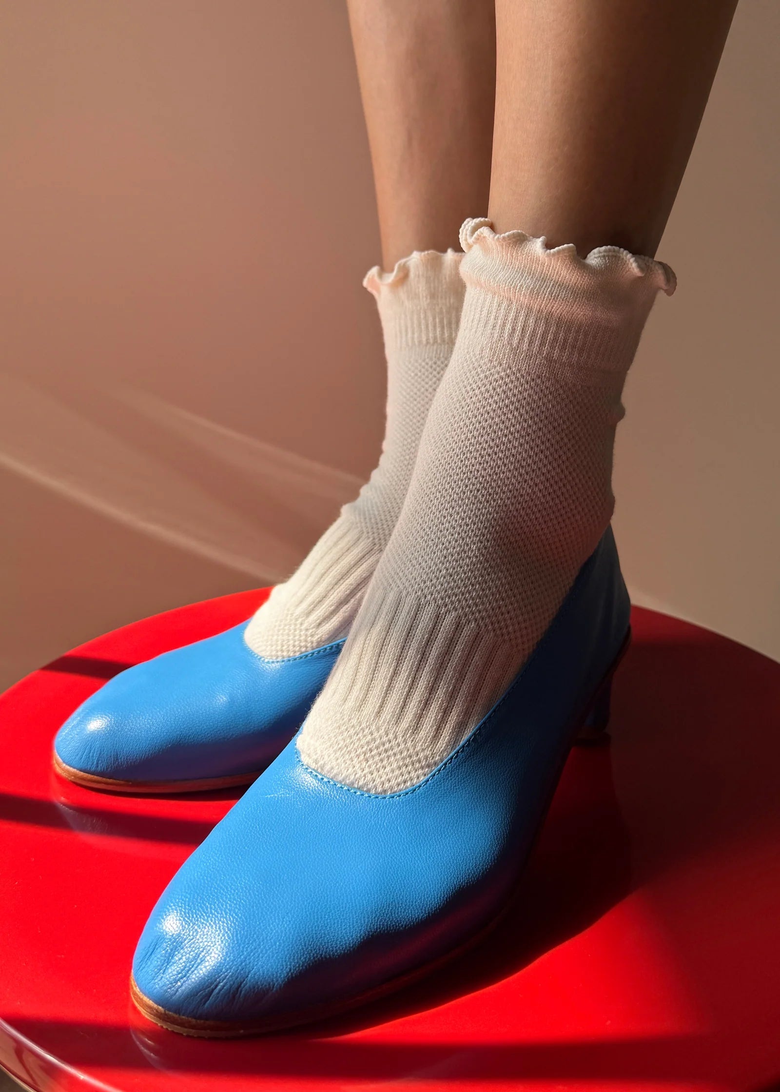 A person wearing white ribbed crew socks with ruffled edges and bright blue leather shoes, standing on a red stool against a warm beige background.