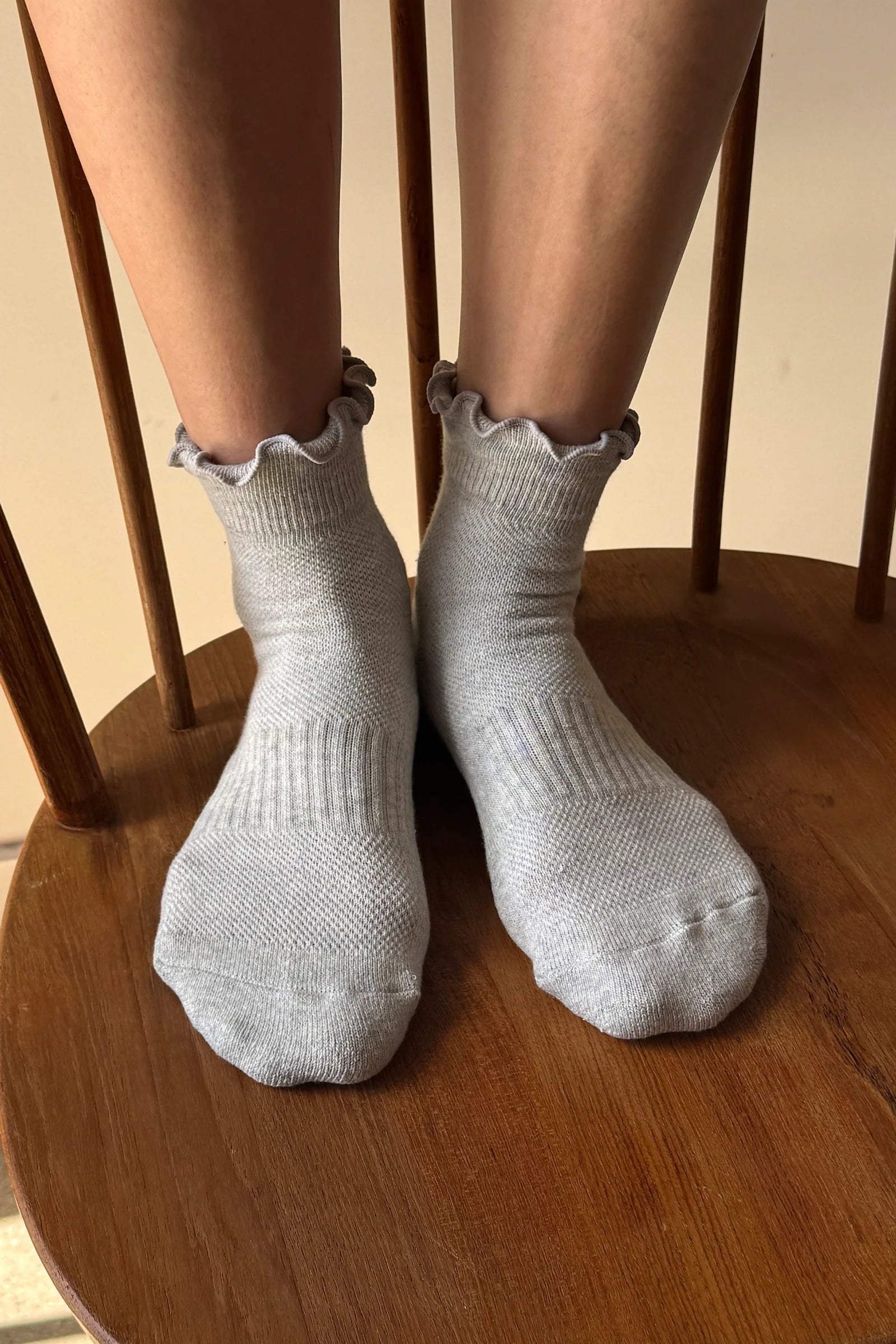 A person wearing light gray ribbed ankle socks with ruffled trim, seated on a wooden chair.