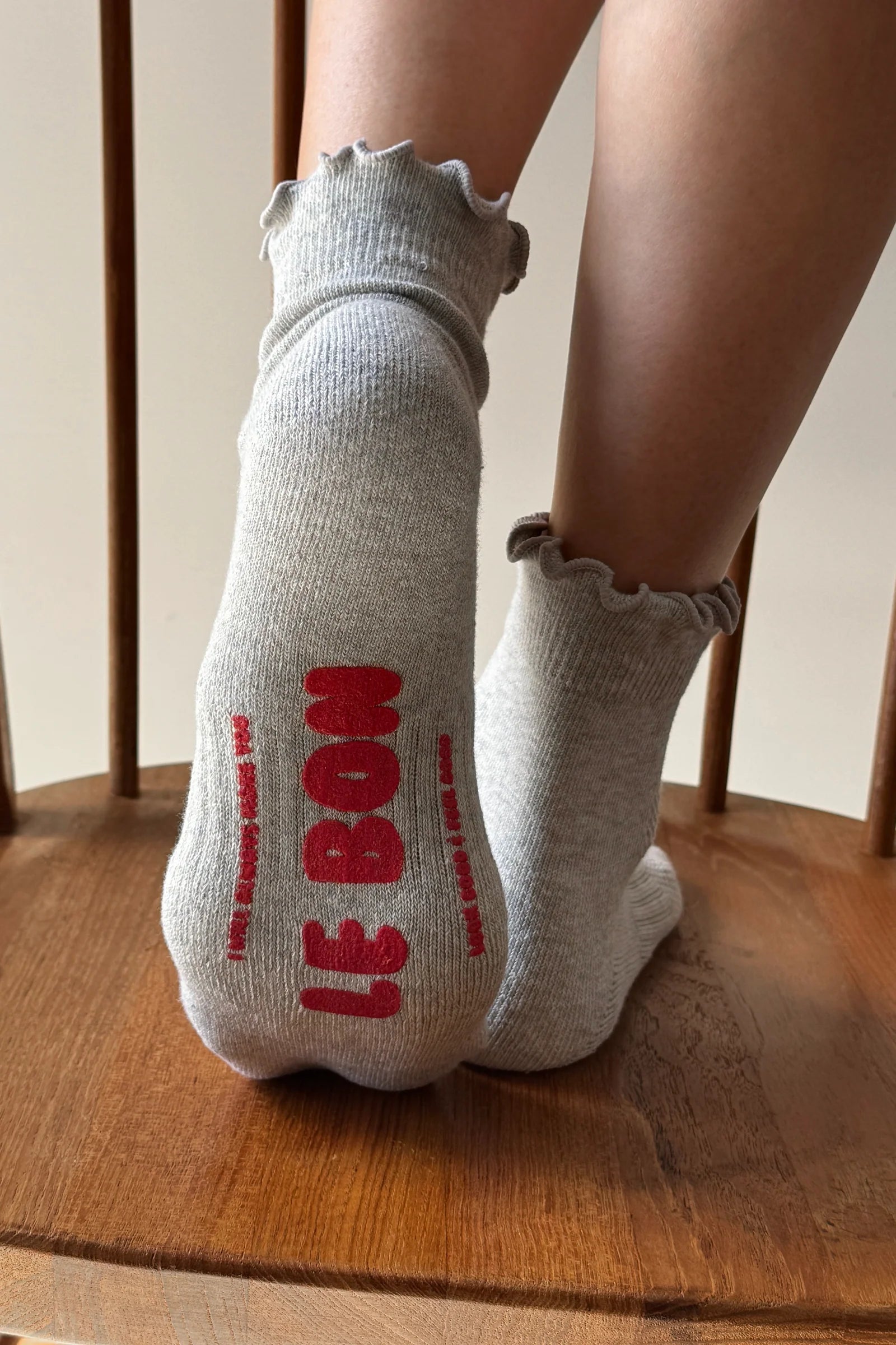 A pair of light gray ankle socks with ruffled edges, worn on a person's feet and standing on a wooden chair. The back of one sock features the text 'LE BON' printed in red.